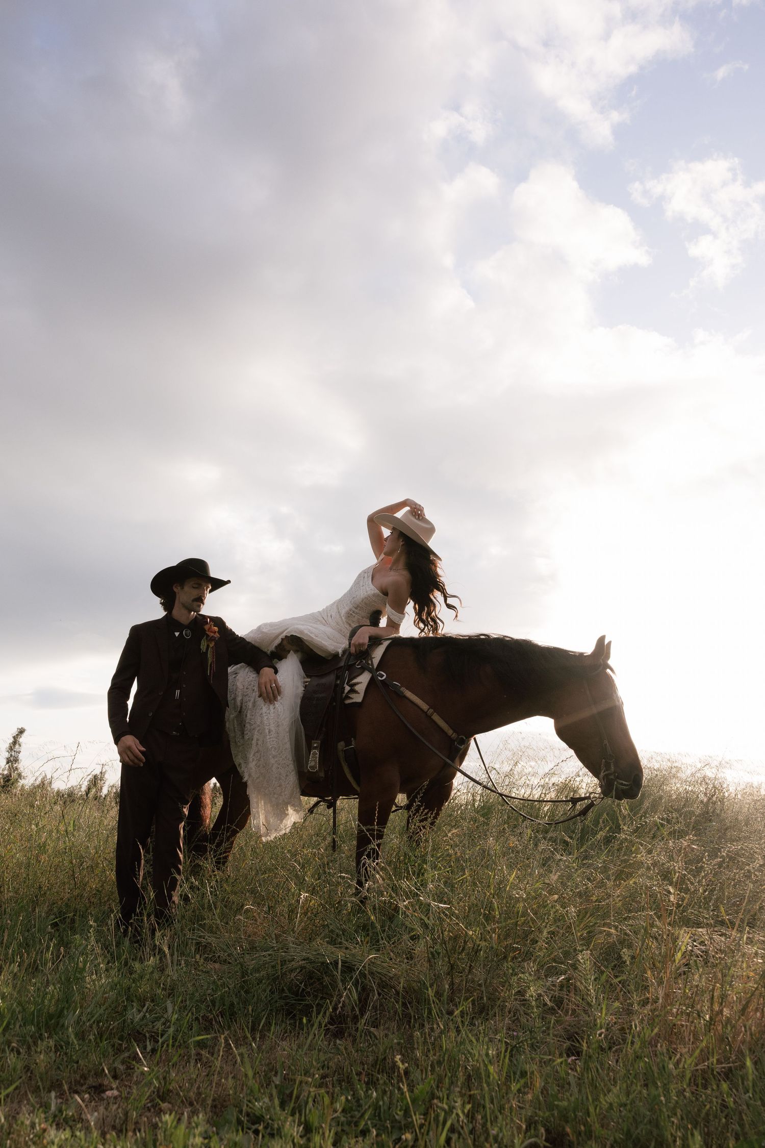 Bride in white dress sitting on horseback with groom in black cowboy hat standing beside her in field.