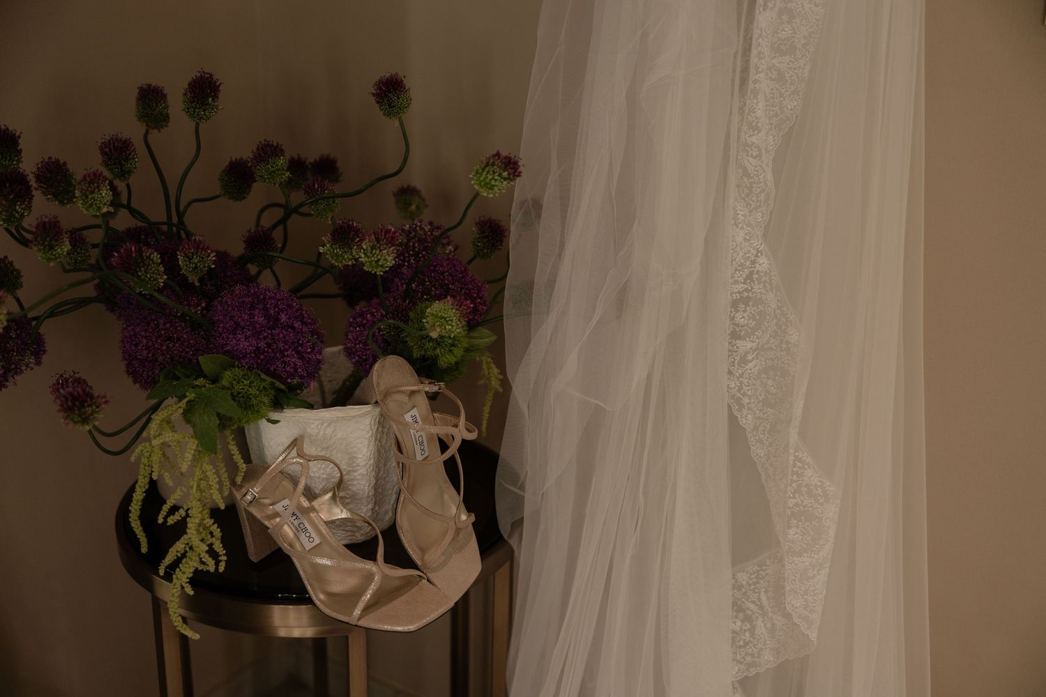 Wedding details flat lay featuring bridal heels, veil, and a purple floral arrangement on a side table.
