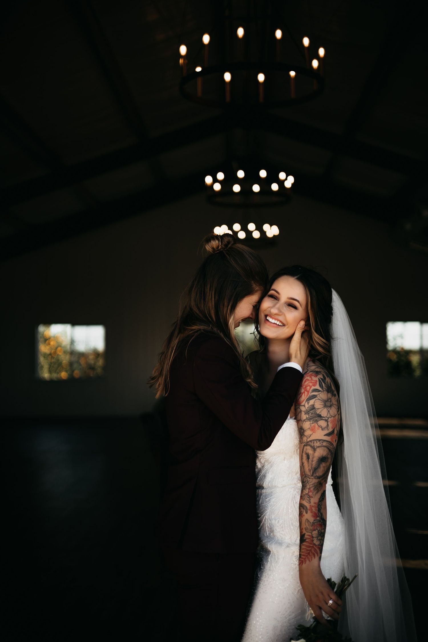 A smiling bride in a lace gown and veil embraces her partner under a candlelit chandelier indoors.