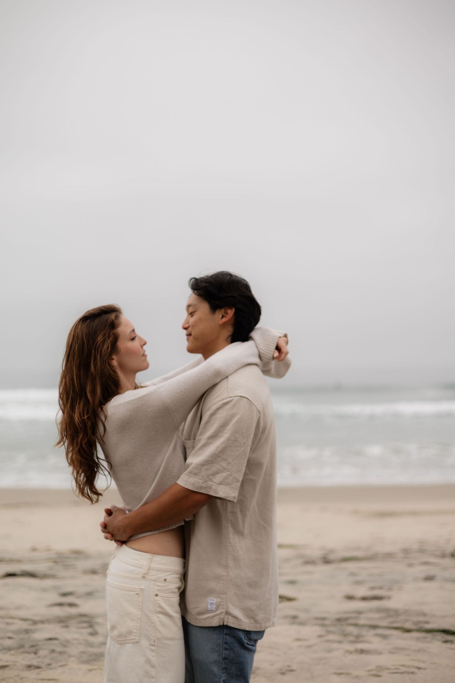 Romantic couple hugging on a sandy beach with ocean waves and overcast sky behind them.