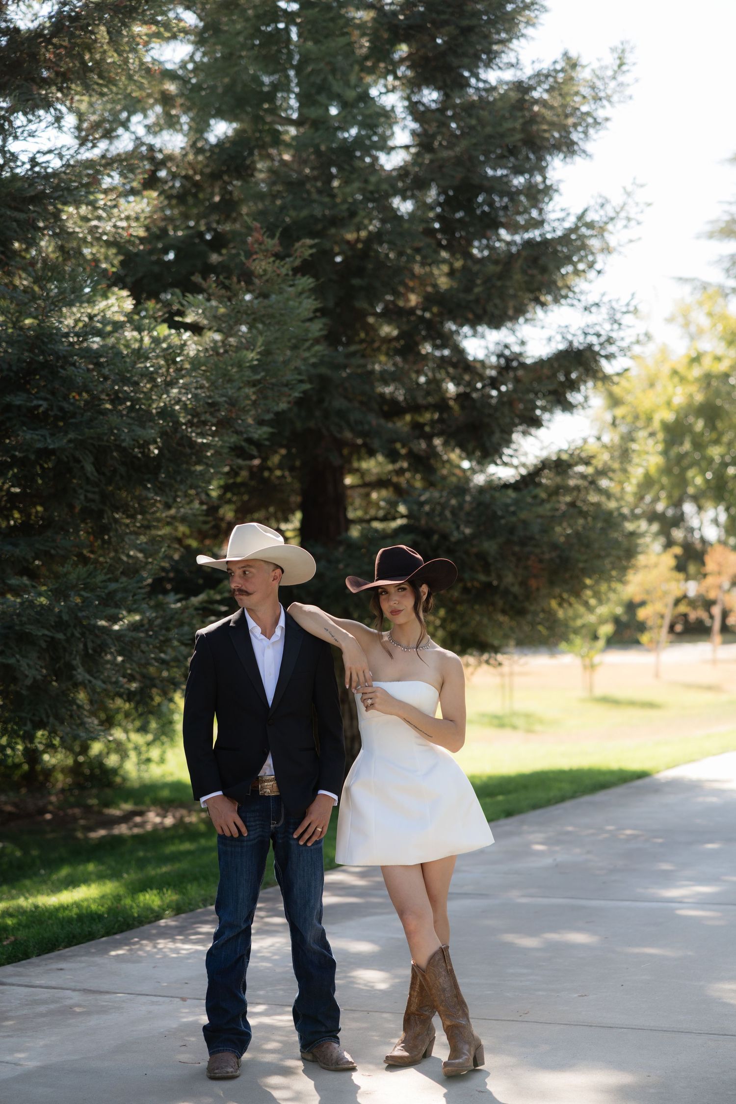 Couple in cowboy hats walking on a path, man in black suit and jeans, woman in white dress and brown boots.