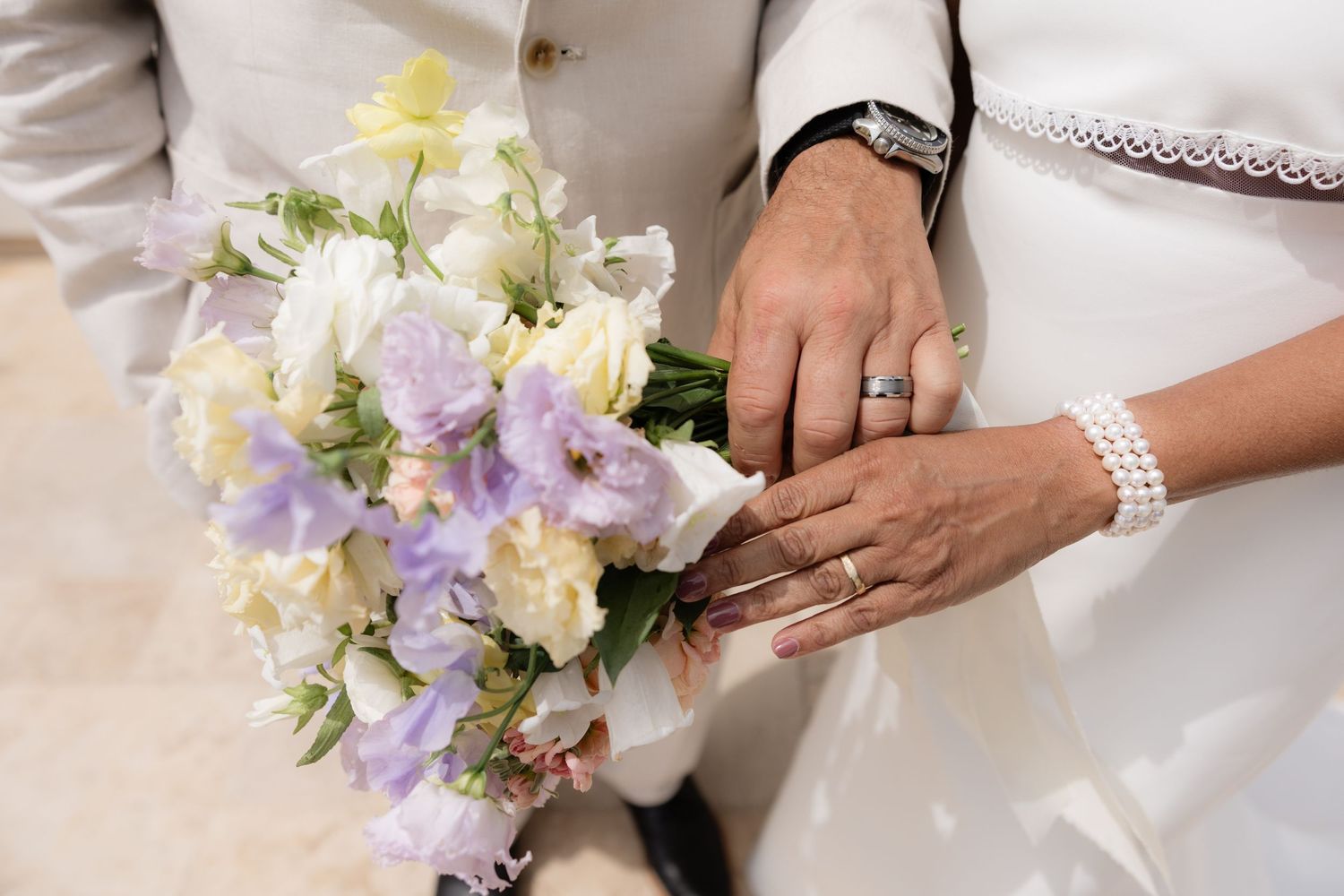 Close-up of newlyweds hands displaying wedding rings beside a pastel bouquet of lavender, white, and yellow flowers.