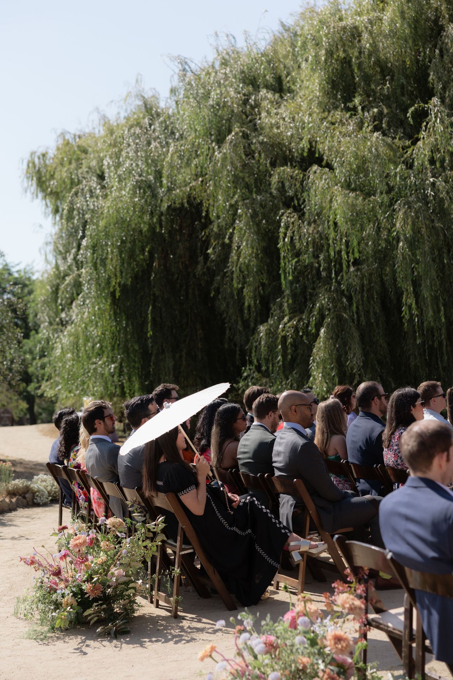 Outdoor wedding ceremony guests seated in wooden chairs under a weeping willow tree, with a woman holding a white parasol.