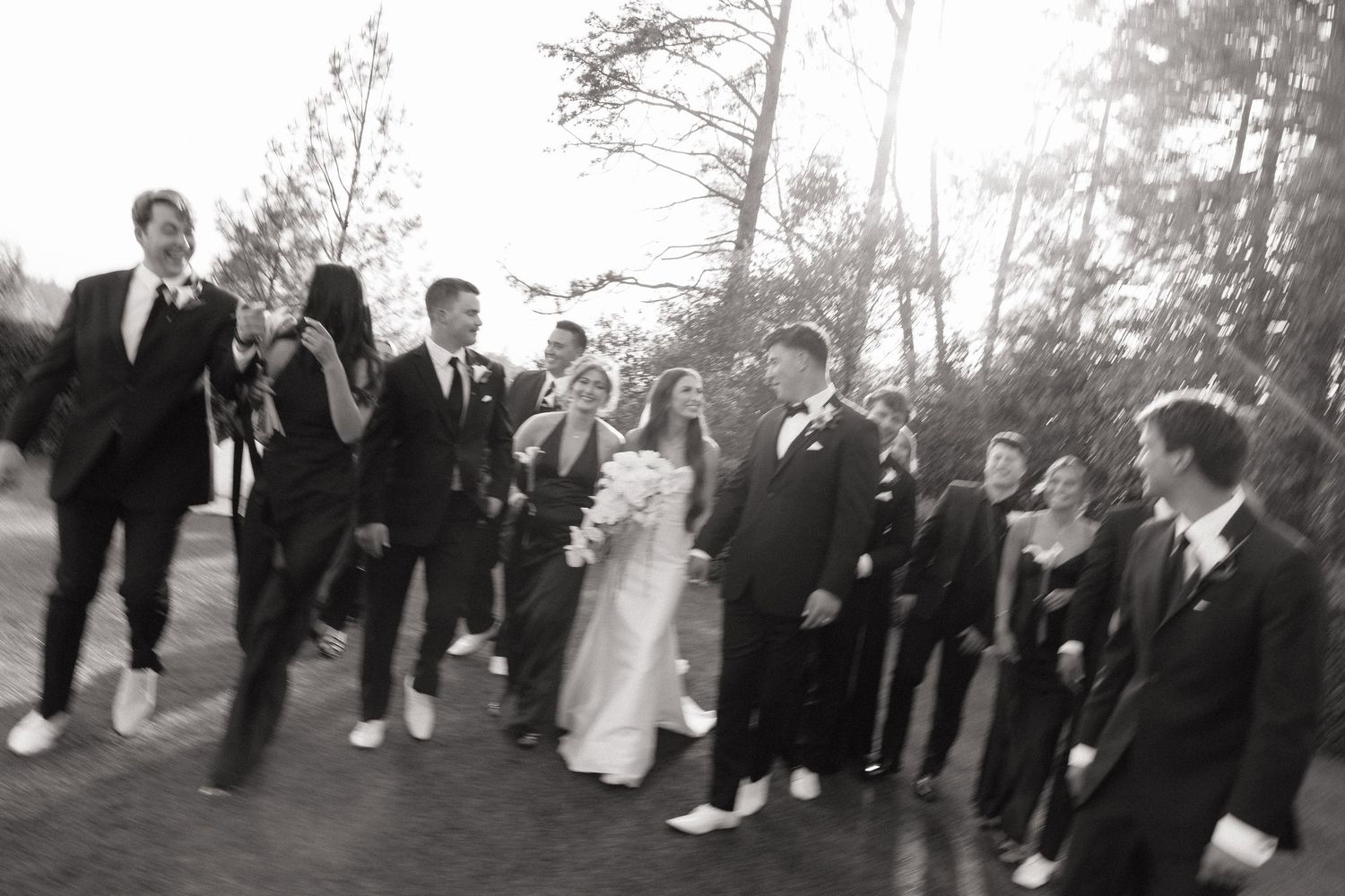 Black and white wedding photo of bride, groom, and wedding party celebrating outdoors among trees.