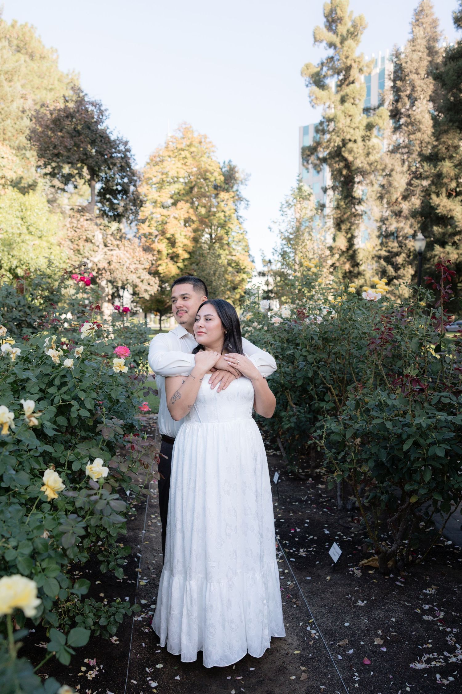 A couple embraces in a lush rose garden, the woman in a white lace gown, surrounded by colorful blooms and trees.