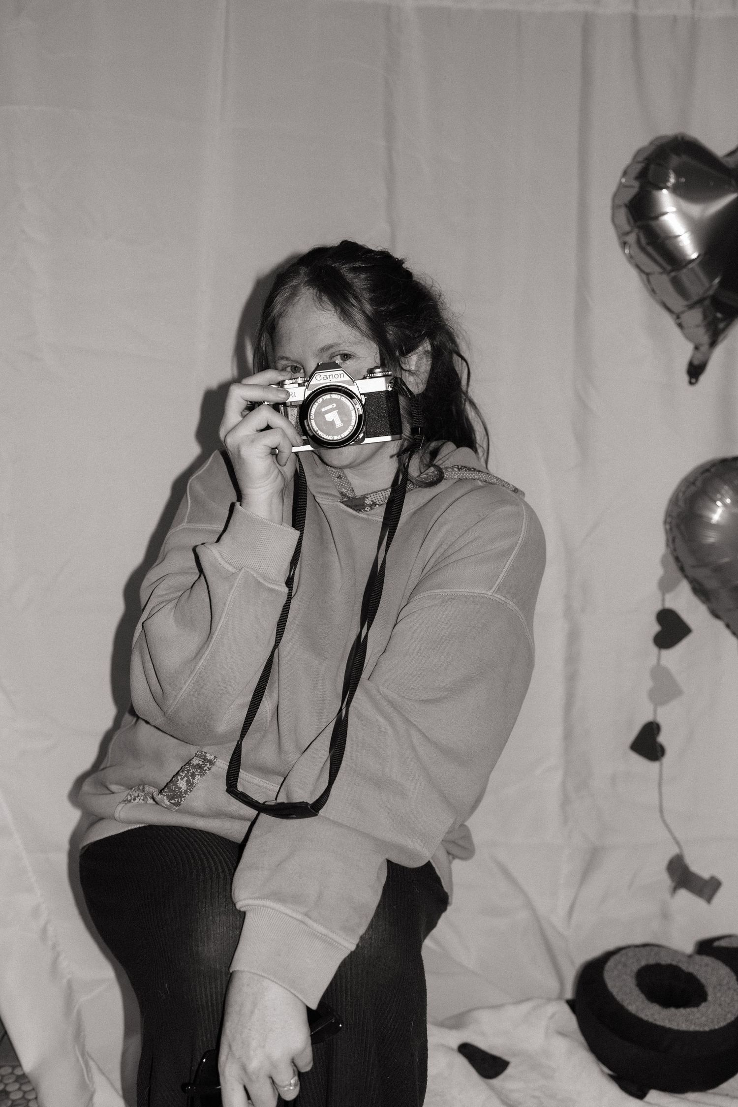 Black and white photo of a woman holding a film camera with heart balloons nearby.