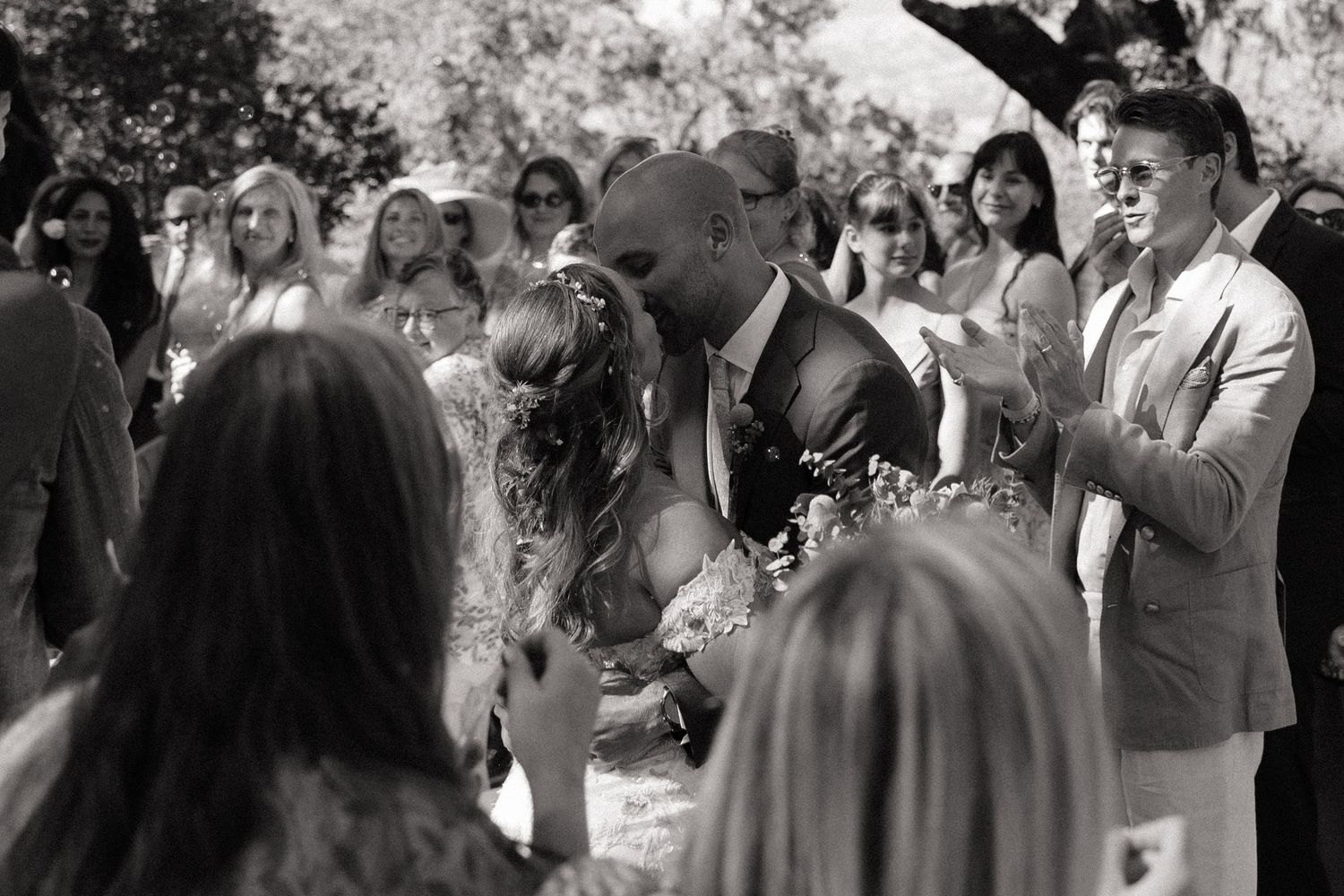 Black and white photo of a couple sharing their first kiss at an outdoor wedding ceremony surrounded by guests.