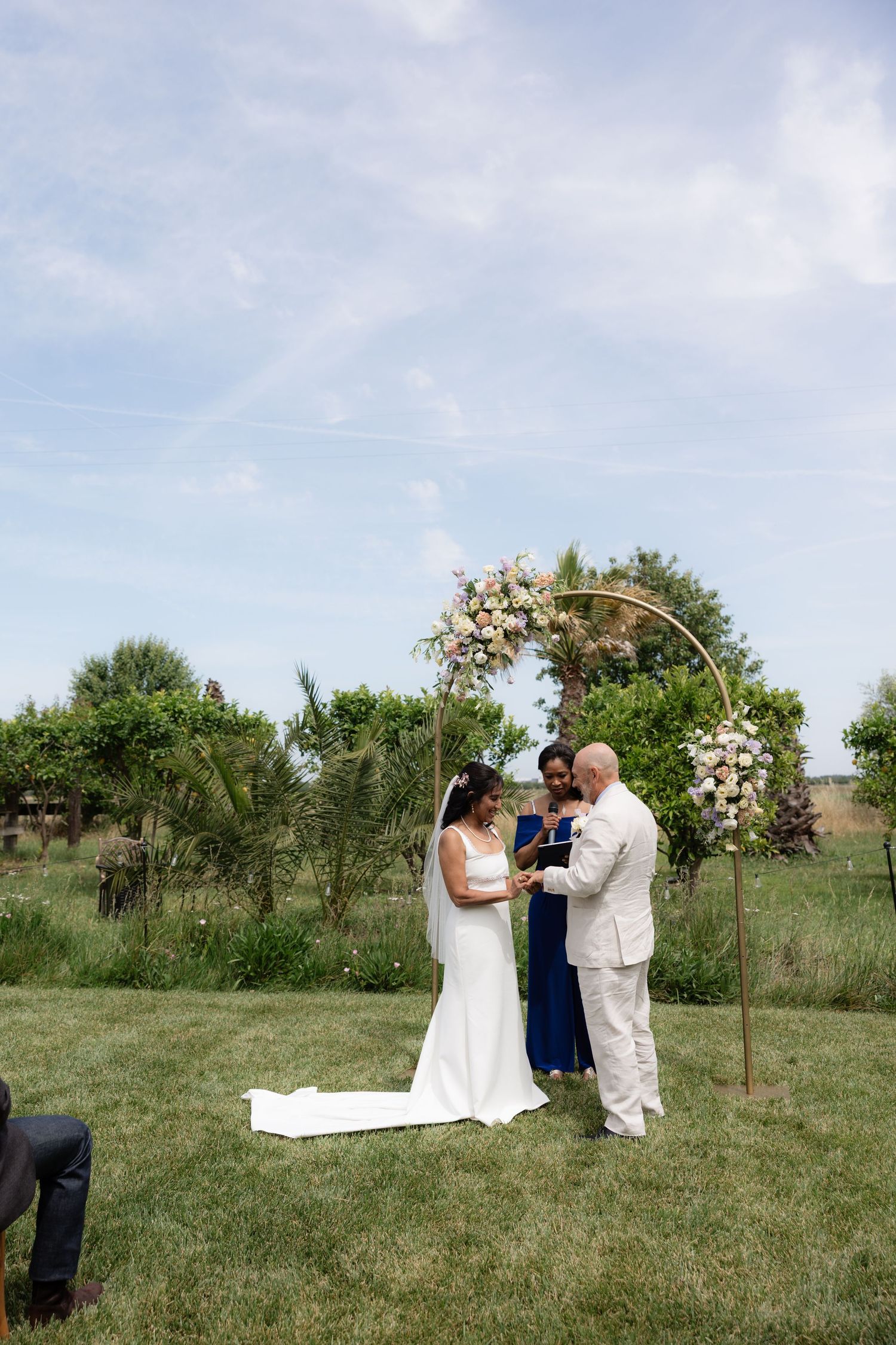Couple exchanges rings at outdoor wedding ceremony beneath a circular gold arch with floral arrangements.