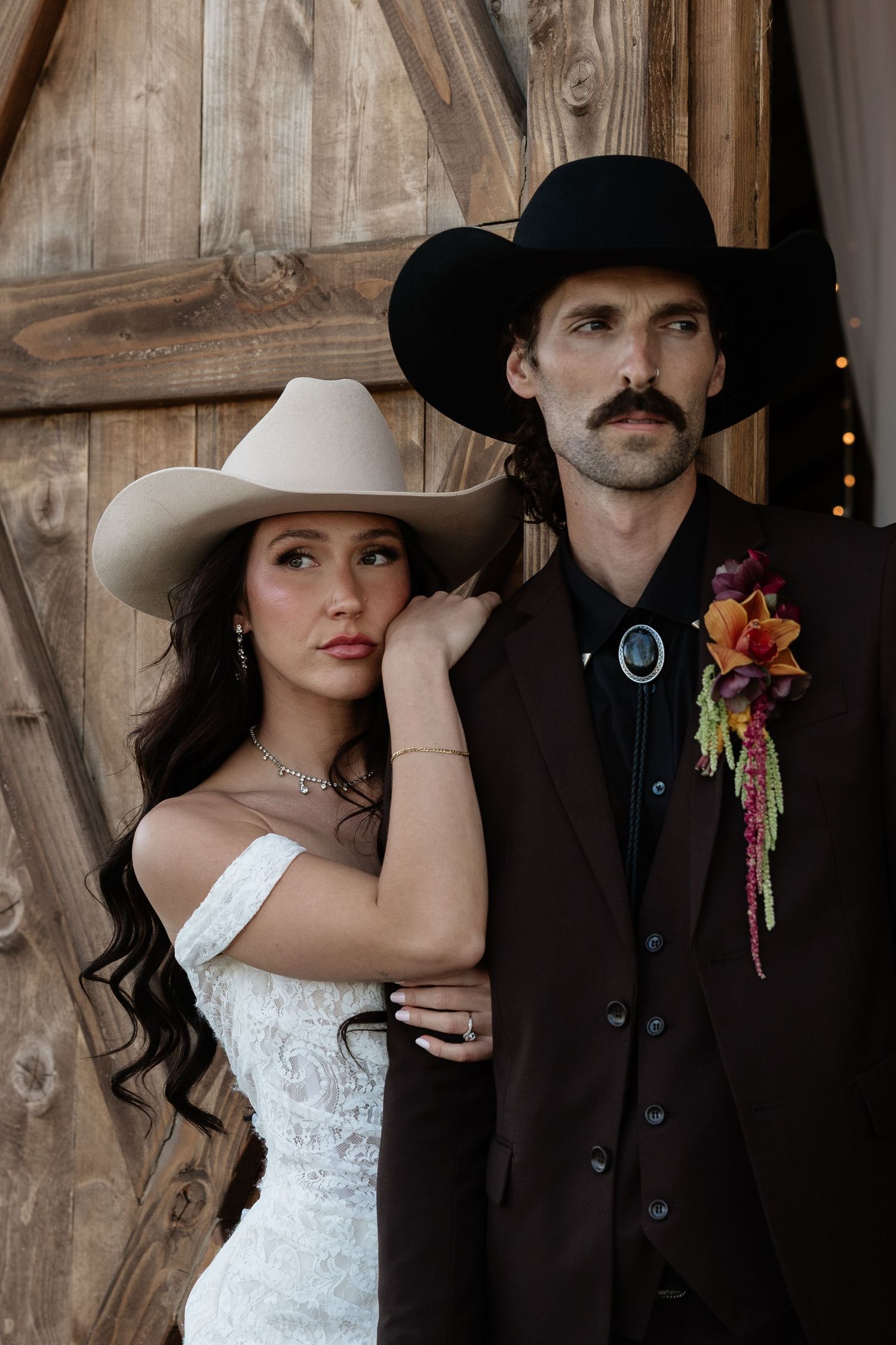 Bride and groom in cowboy hats pose before rustic wooden barn doors, groom wearing floral boutonniere.