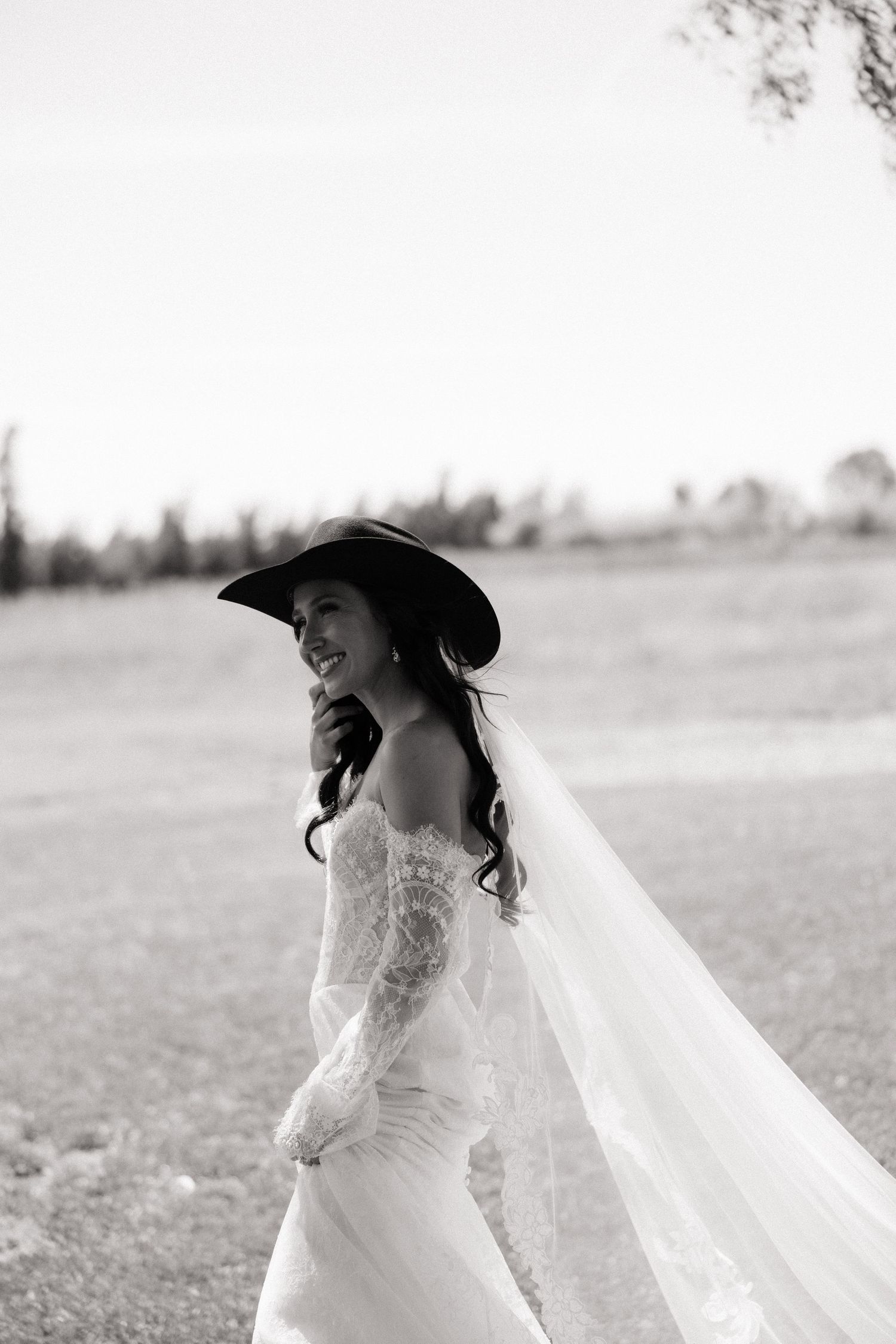 Smiling bride in white lace gown and black cowboy hat with veil flowing in the breeze.