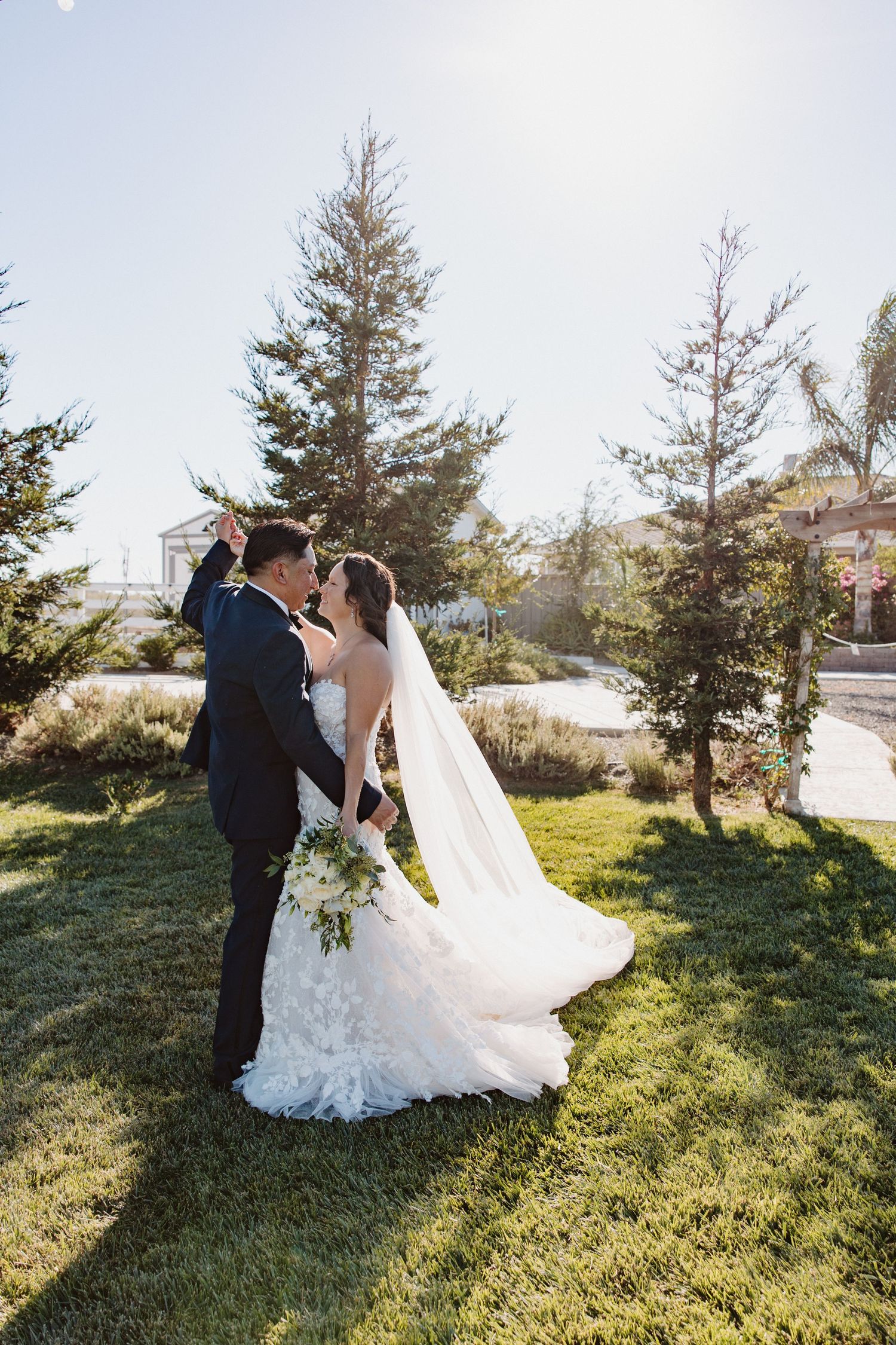 Bride and groom share a tender kiss on a sunny lawn with veil flowing during wedding portraits.