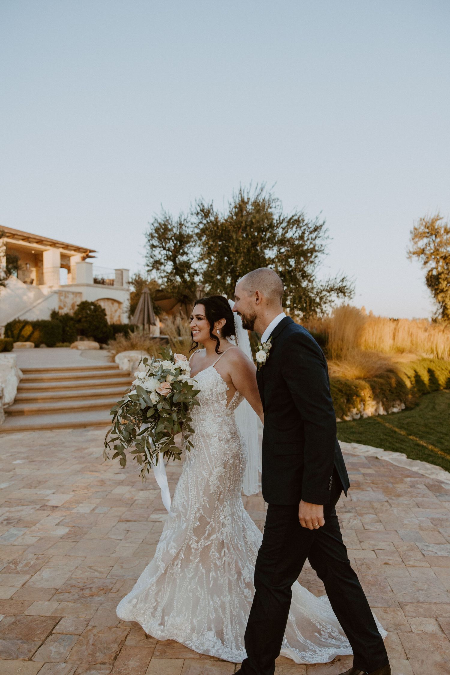 Bride and groom walk together smiling at sunset outside elegant stone wedding venue.