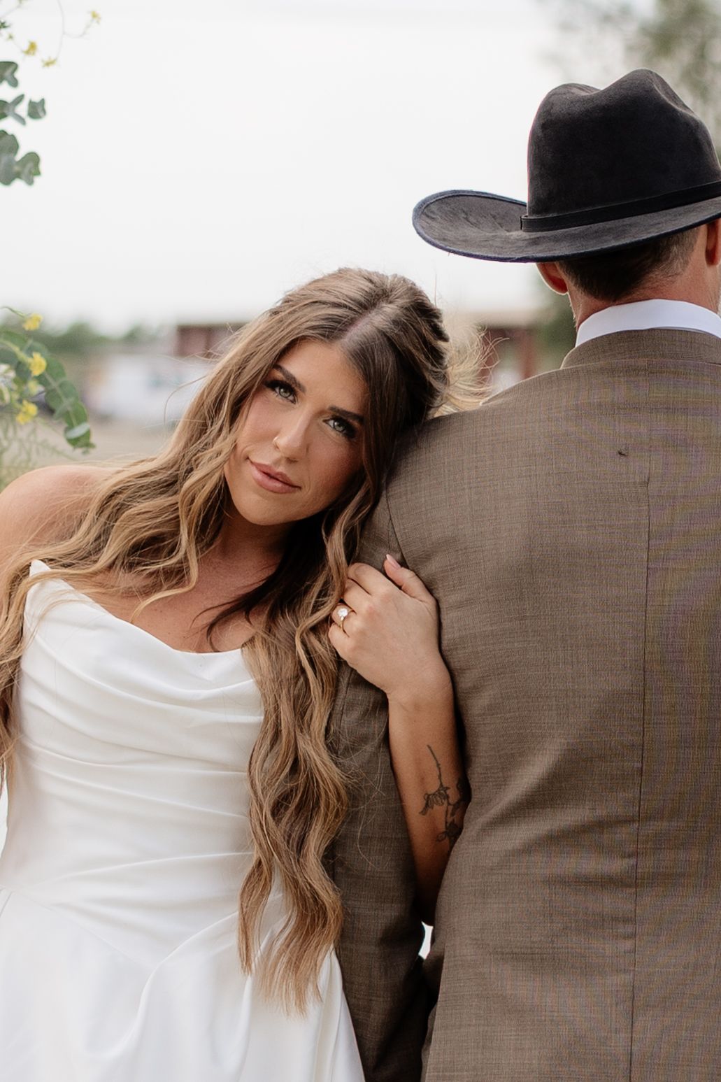 Bride in white gown leans on groom's shoulder, looking back at camera during outdoor wedding photo session.