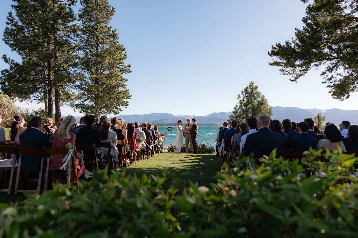 Outdoor lakeside wedding ceremony with guests seated on lawn overlooking stunning blue mountain lake view.