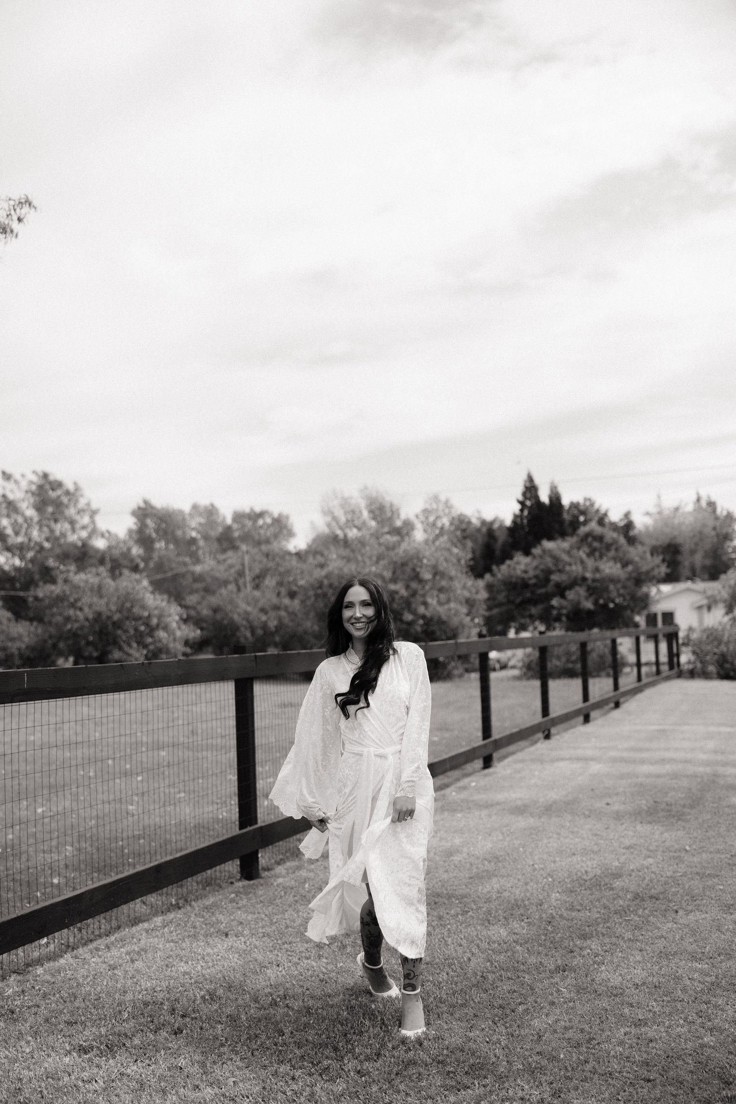 Bride in flowing white dress walking along a black fence on a grassy outdoor wedding venue.