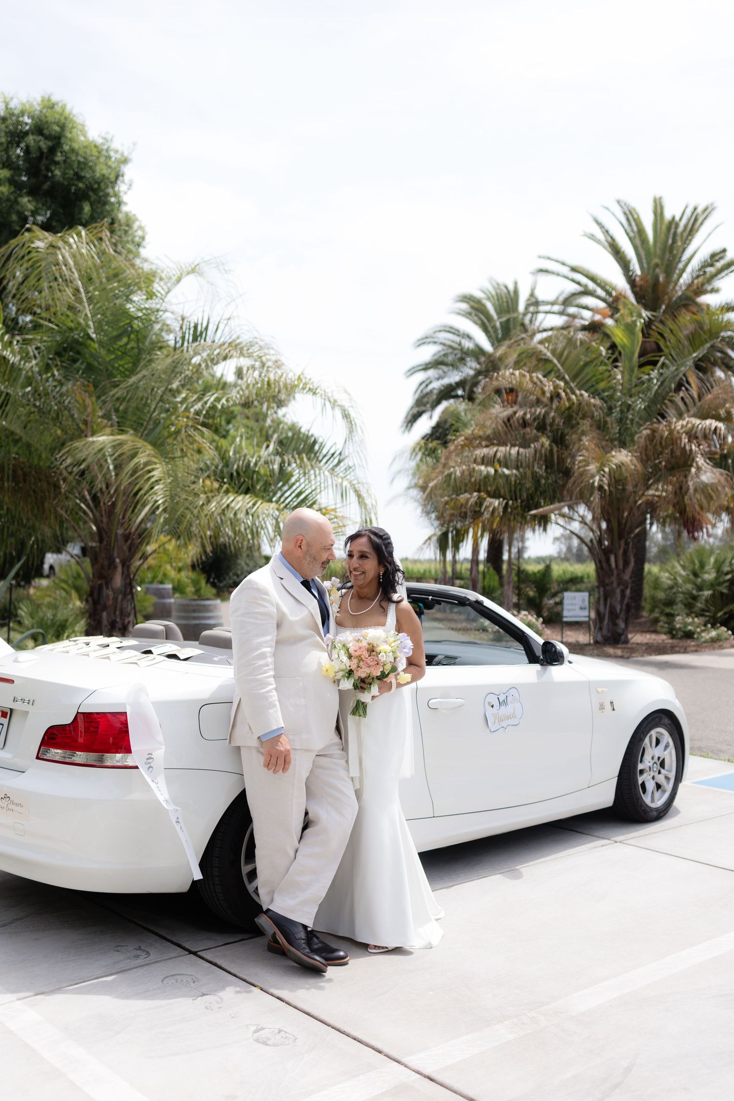 Bride and groom pose together beside a white convertible car surrounded by tall palm trees.
