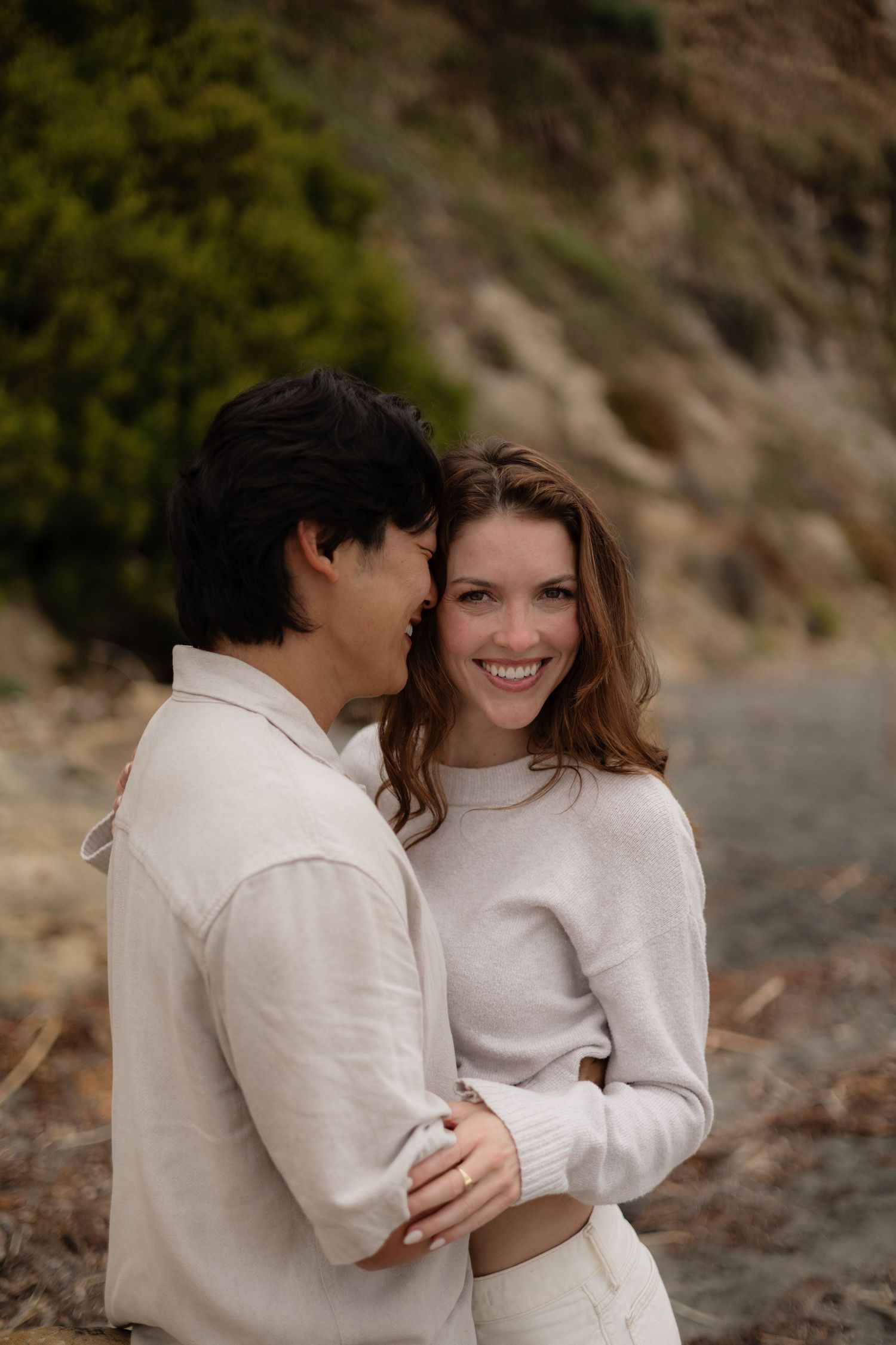 A smiling couple in matching cream outfits embrace outdoors near a rocky hillside with lush greenery.
