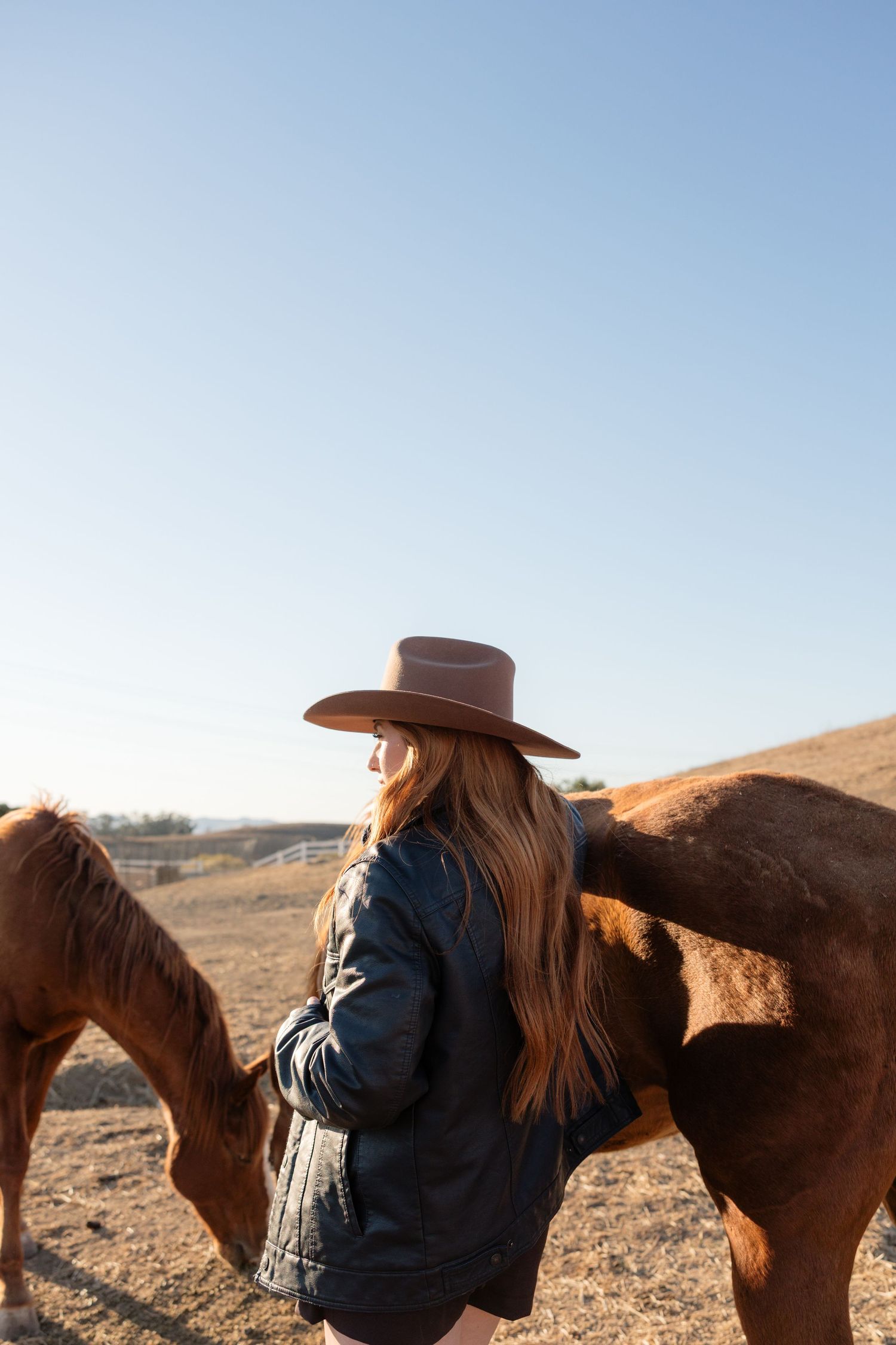 Woman in cowboy hat and denim jacket standing with brown horses on a sunny ranch.