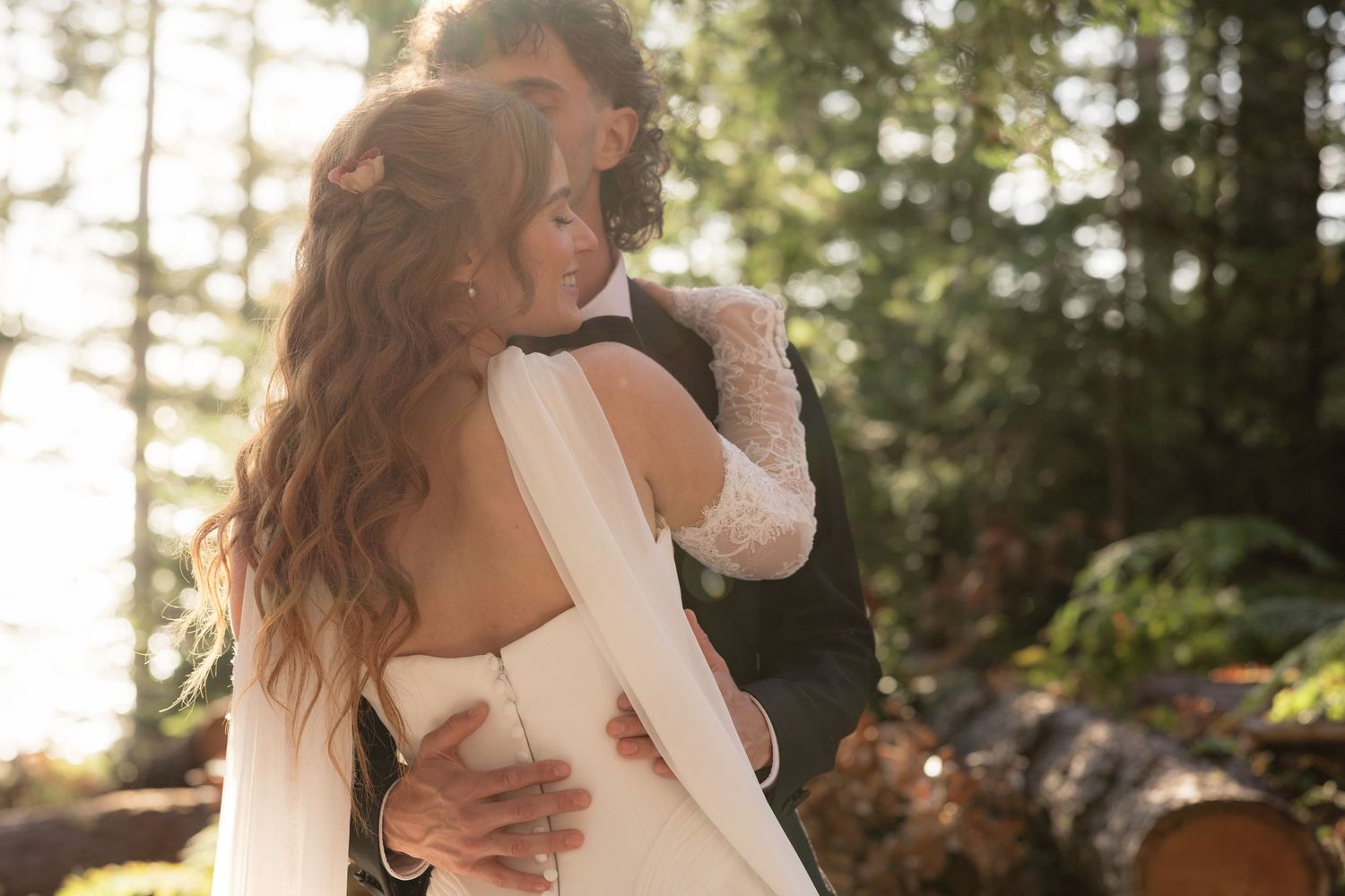 A bride and groom embrace romantically in a sunlit forest, the bride wearing a backless white gown with flowing veil.