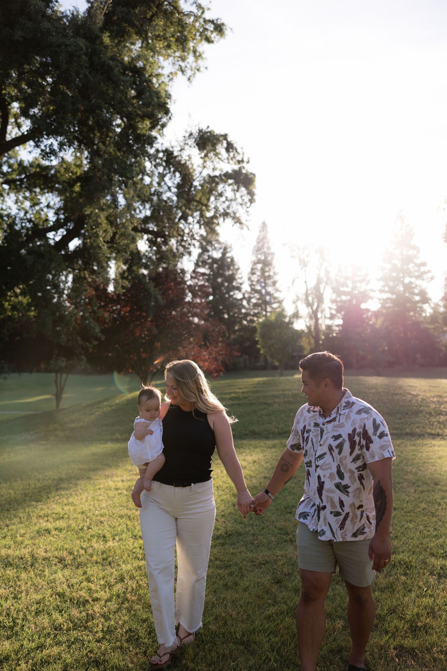Happy family of three walking hand in hand through a sunlit park, mother carrying their infant baby.