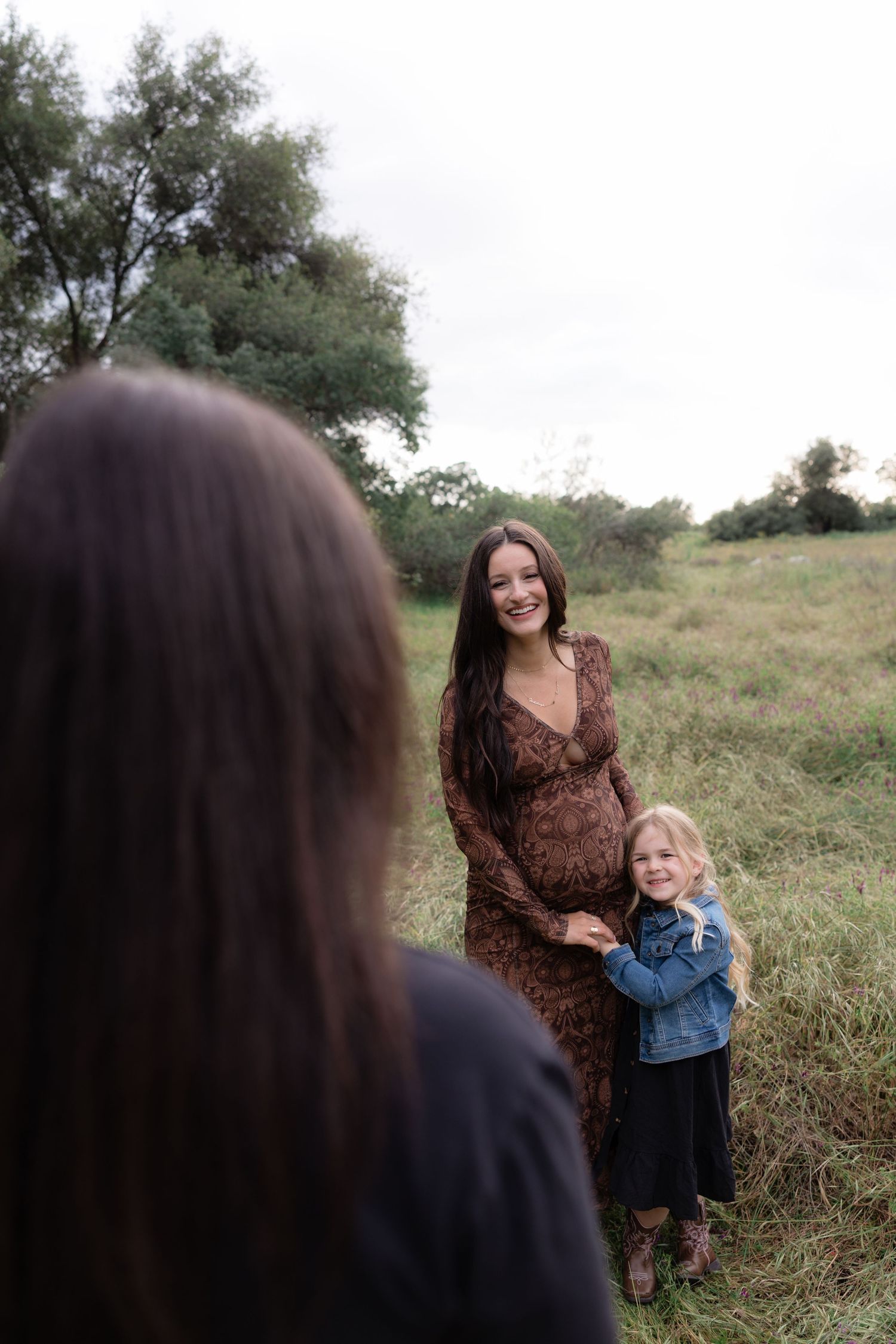 Smiling pregnant woman in brown lace dress with young girl in field during outdoor maternity photoshoot.