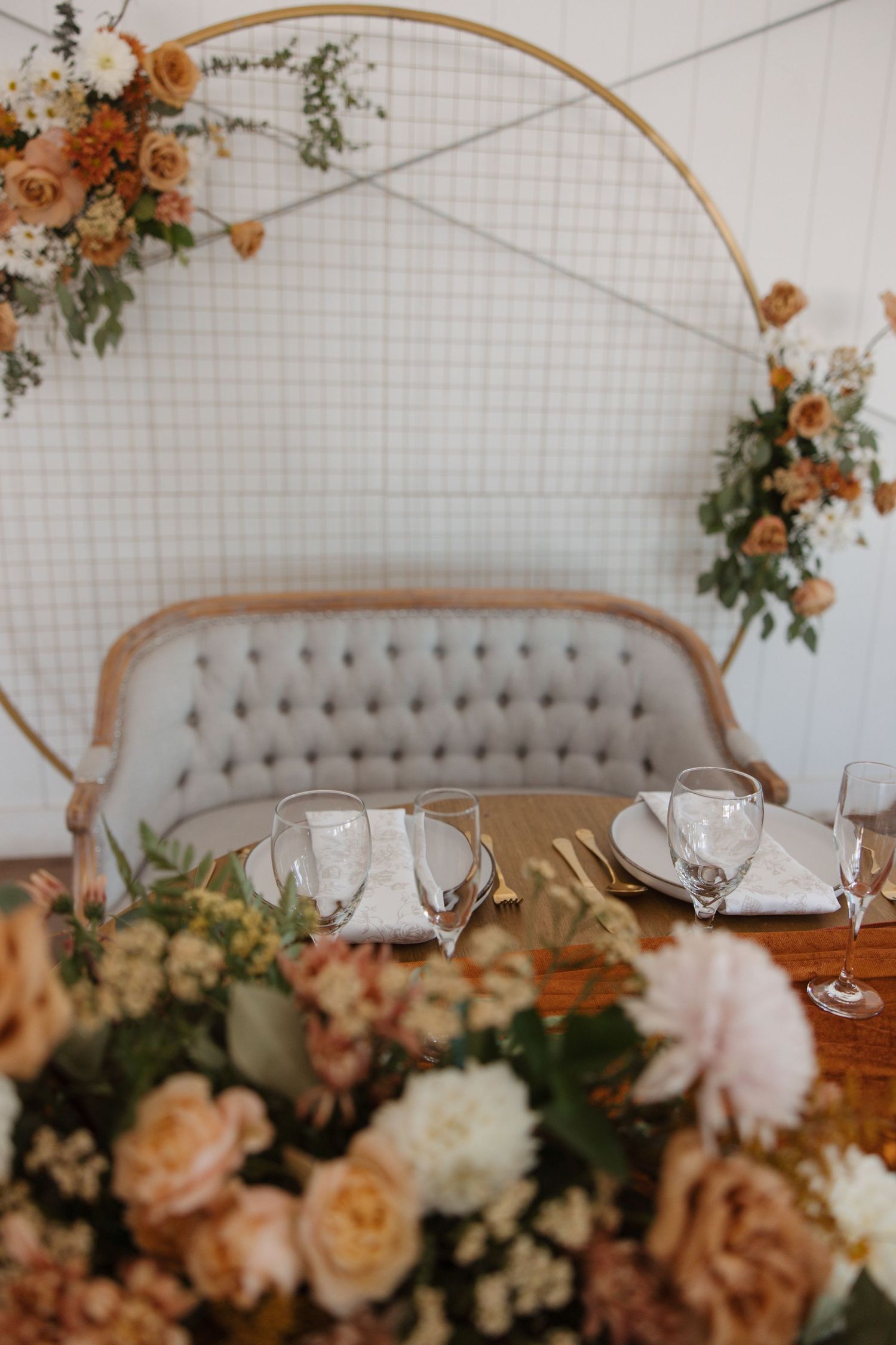 Elegant sweetheart table with tufted loveseat, floral arrangements, and gold circular arch backdrop.