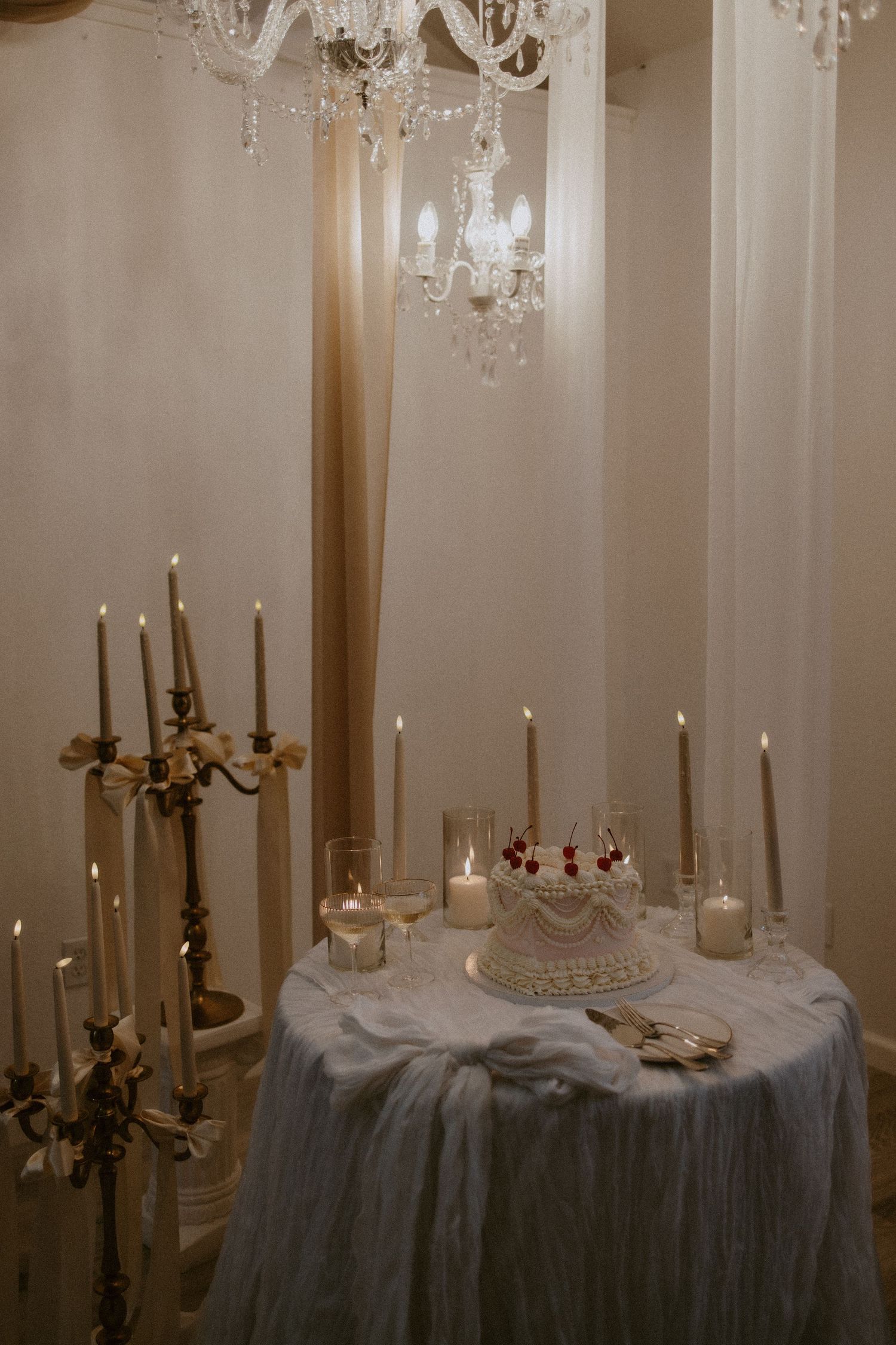 Romantic wedding cake table with candles and crystal chandelier in elegant white room.