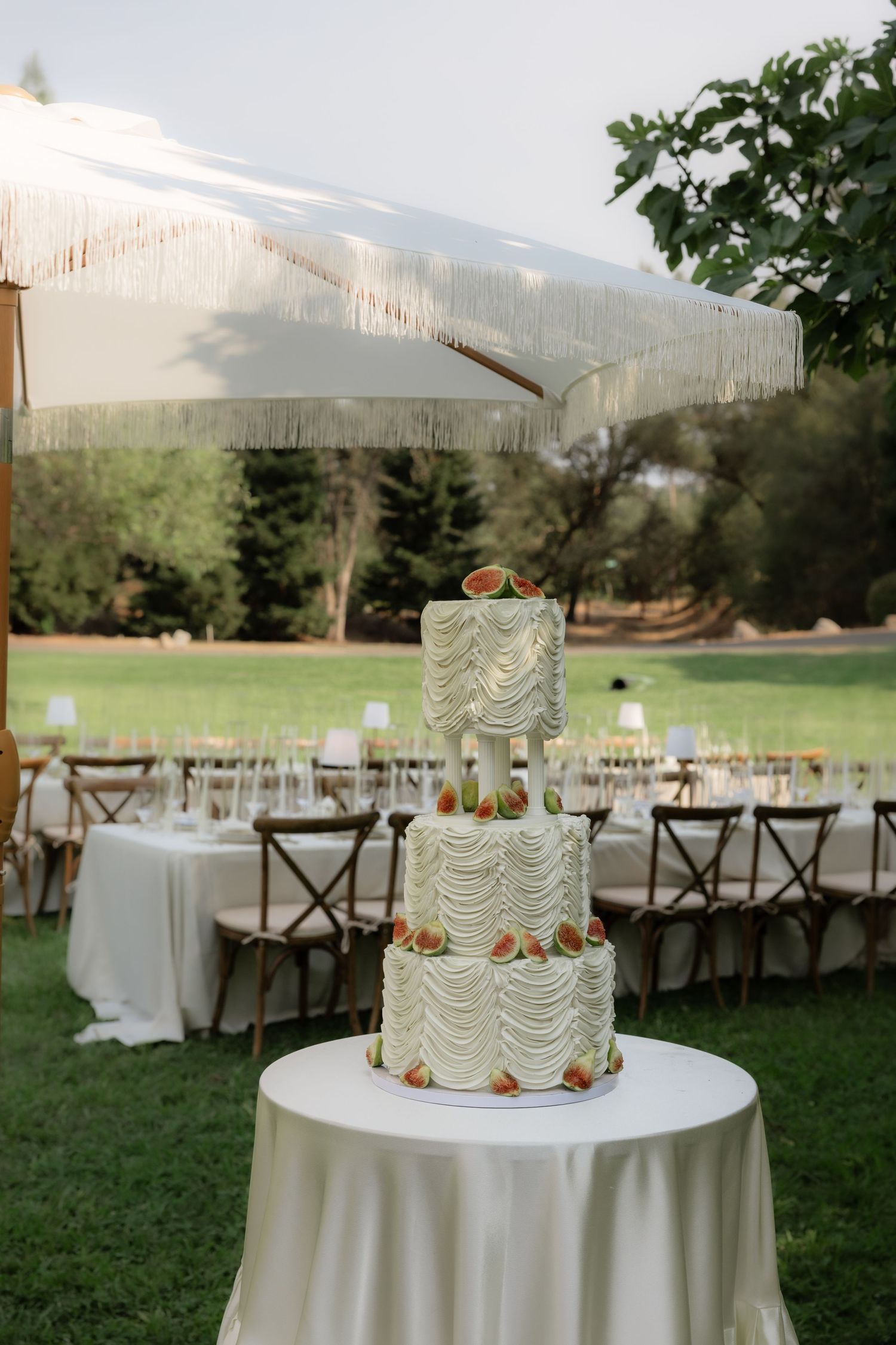 Elegant three-tier white wedding cake with ruffled frosting and fig decorations displayed outdoors under a fringed umbrella.