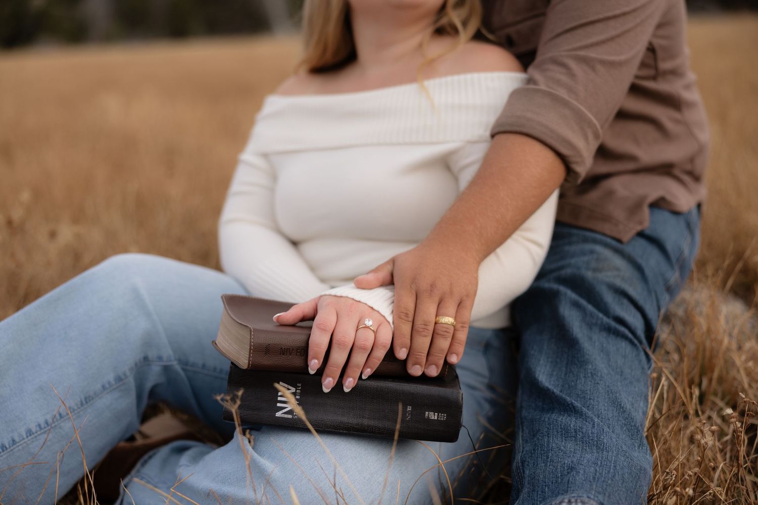 Couple sitting in golden field, woman in white off-shoulder sweater holding Bible with engagement ring visible.