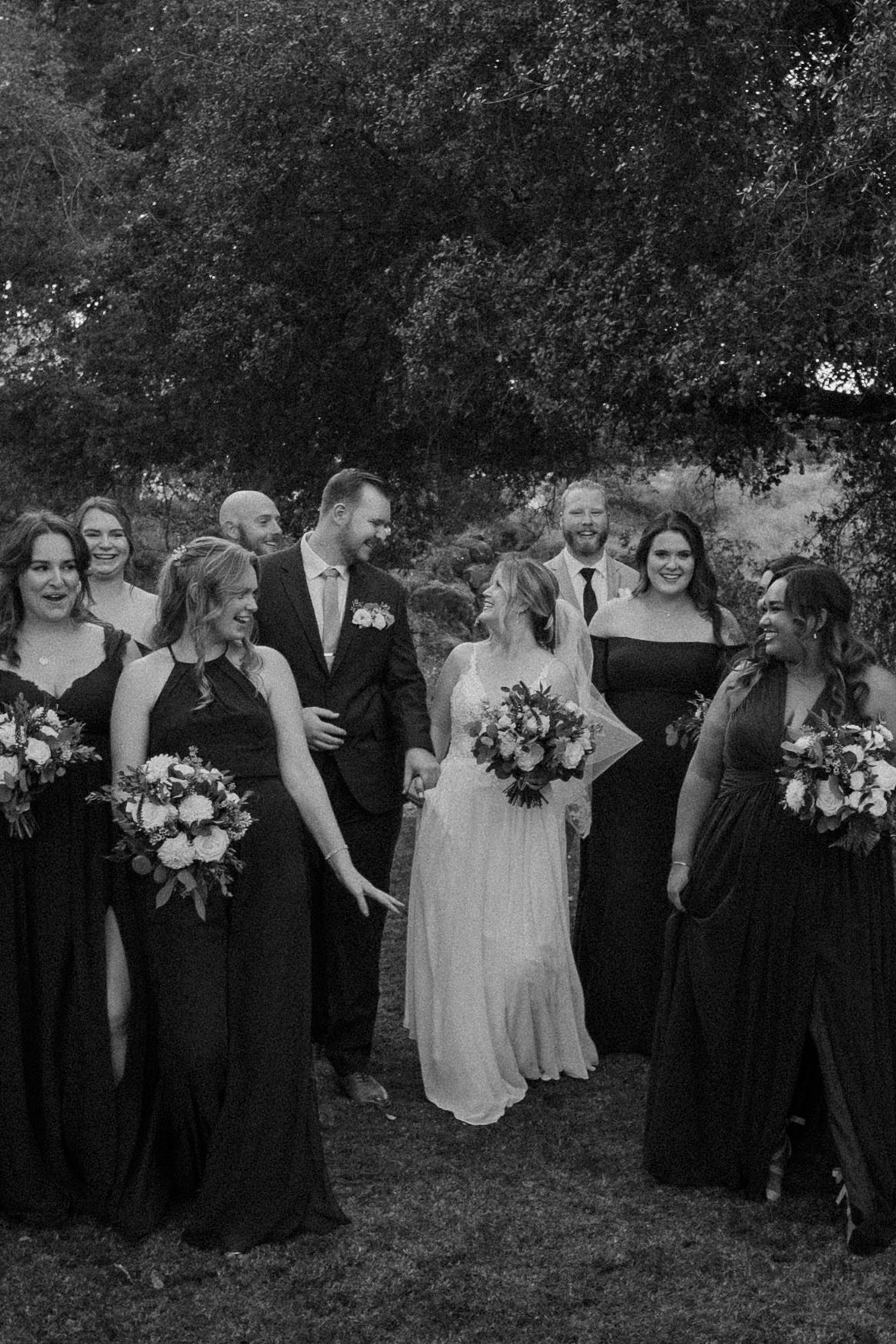 Black and white photo of bride and groom walking with wedding party in dark formal attire outdoors.