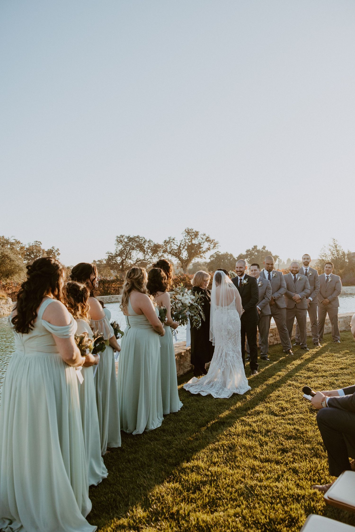 Outdoor wedding ceremony with bridesmaids in sage green gowns and groomsmen in gray suits.