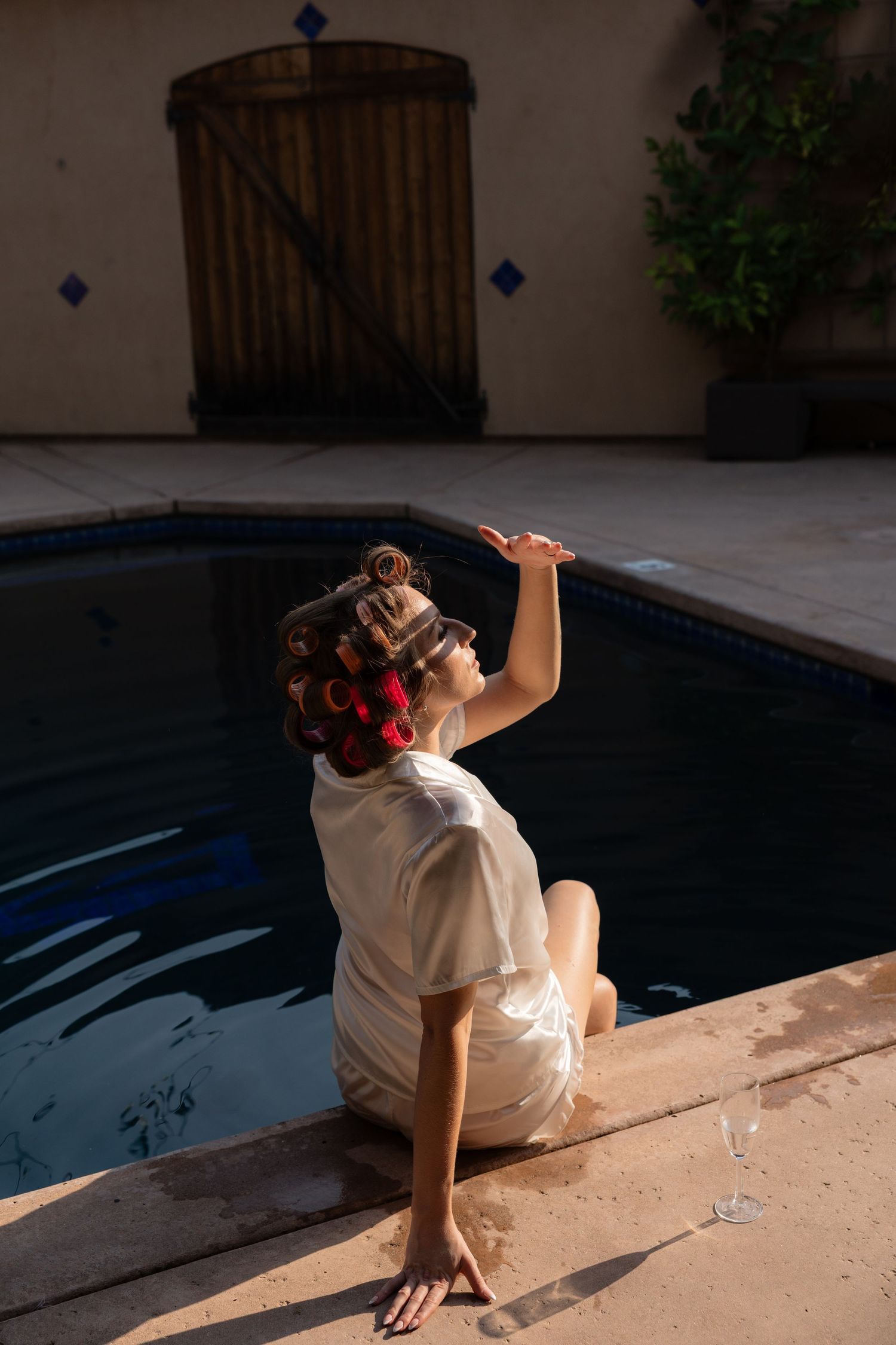 Woman in white robe with hair curlers sitting by a pool edge, reaching up in a sunlit courtyard.