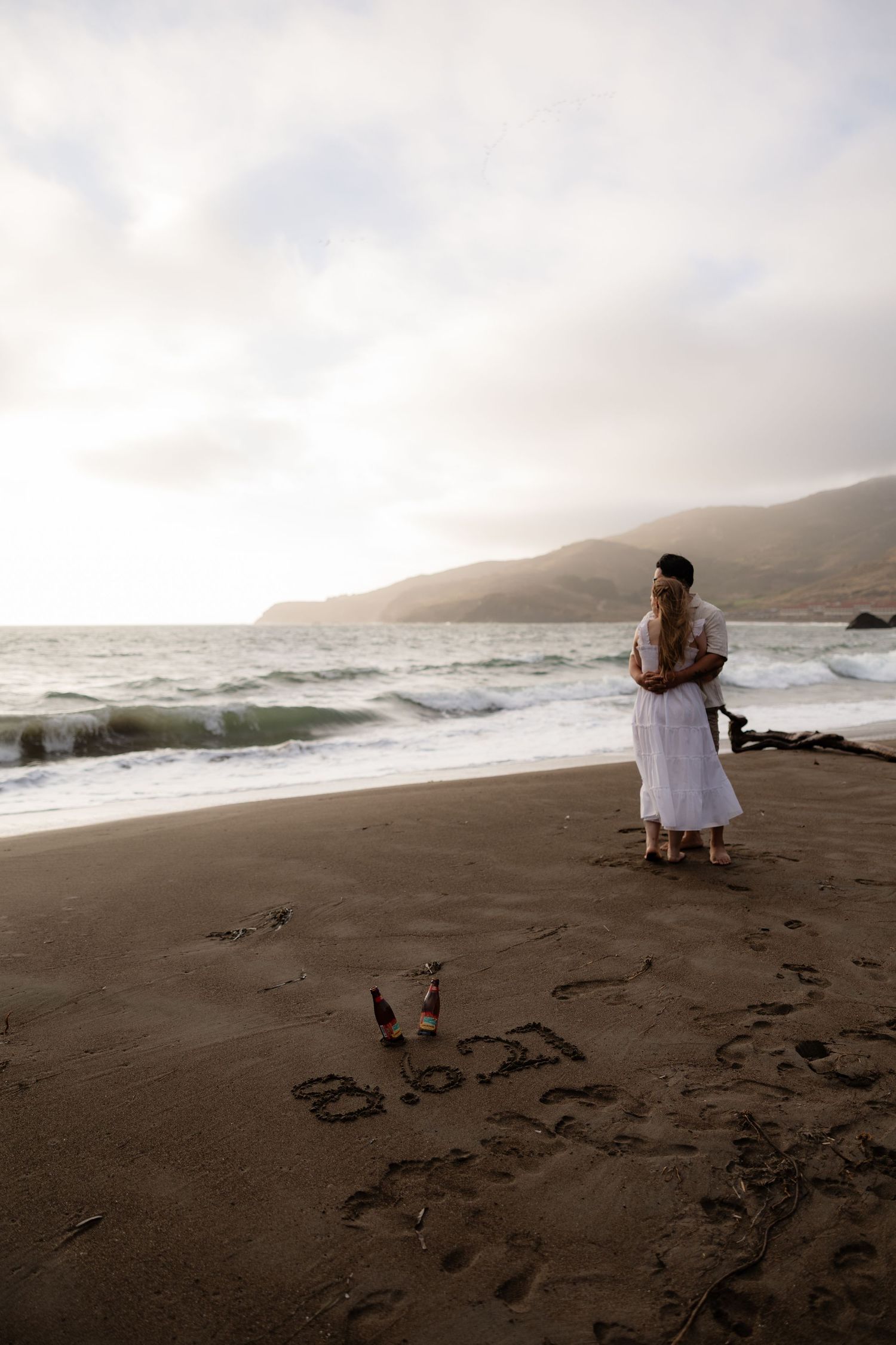 Couple embracing on a misty beach at sunset with words written in sand and ocean waves in the background.