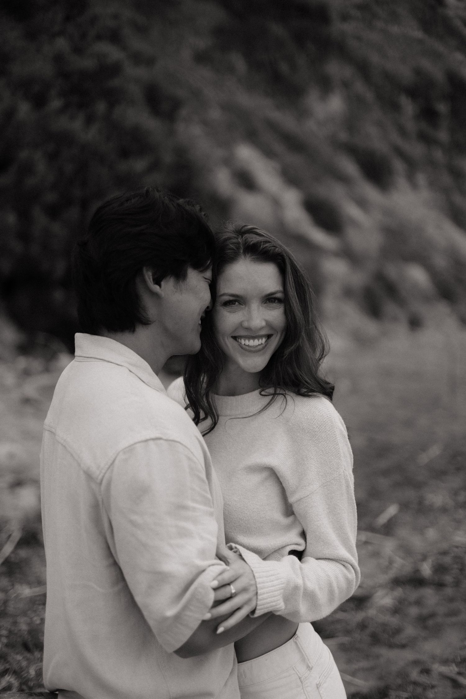 Black and white photo of a smiling couple embracing outdoors, man whispering to woman in cozy sweaters.