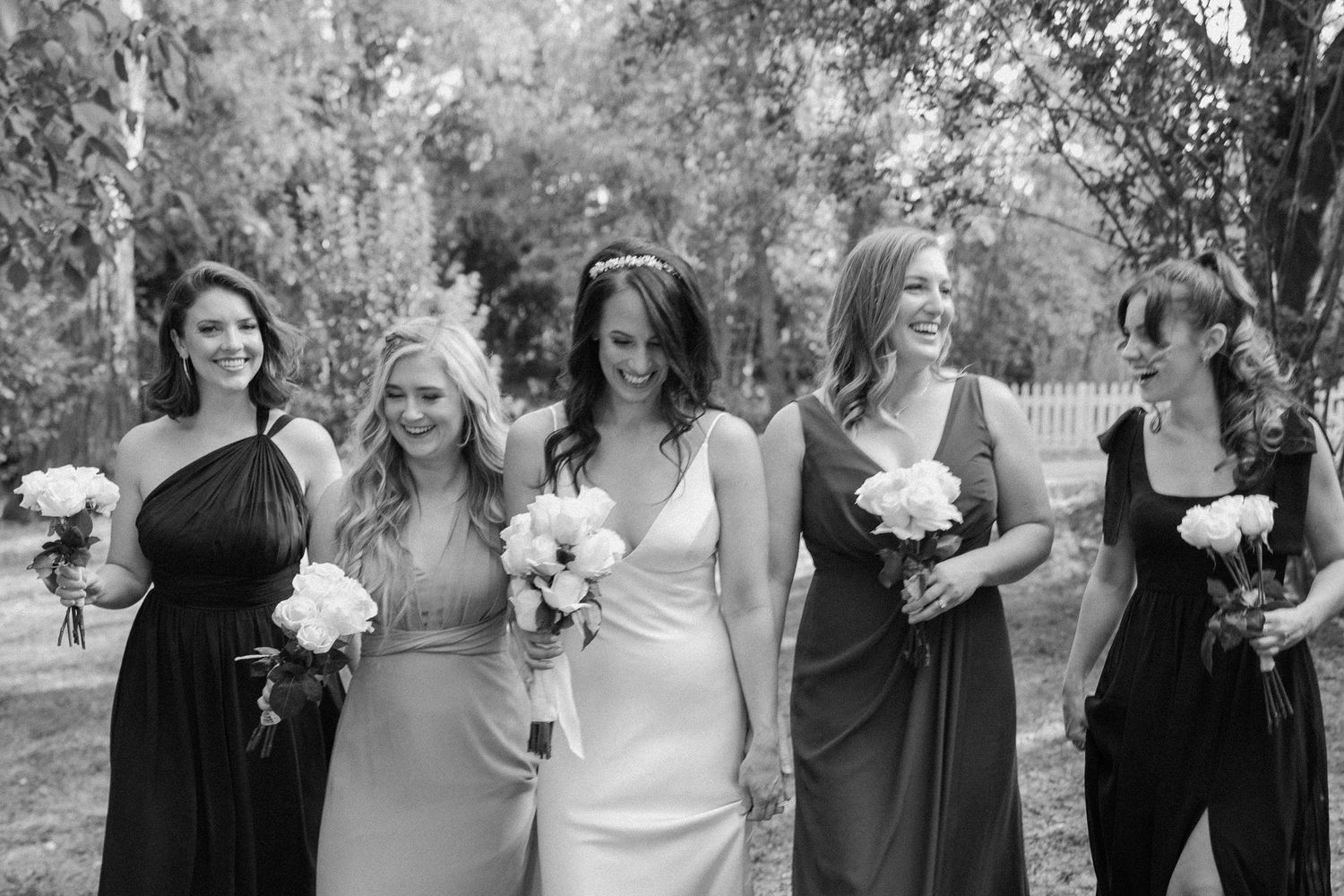 Bride and bridesmaids in mismatched dark dresses laughing together outdoors in black and white photo.