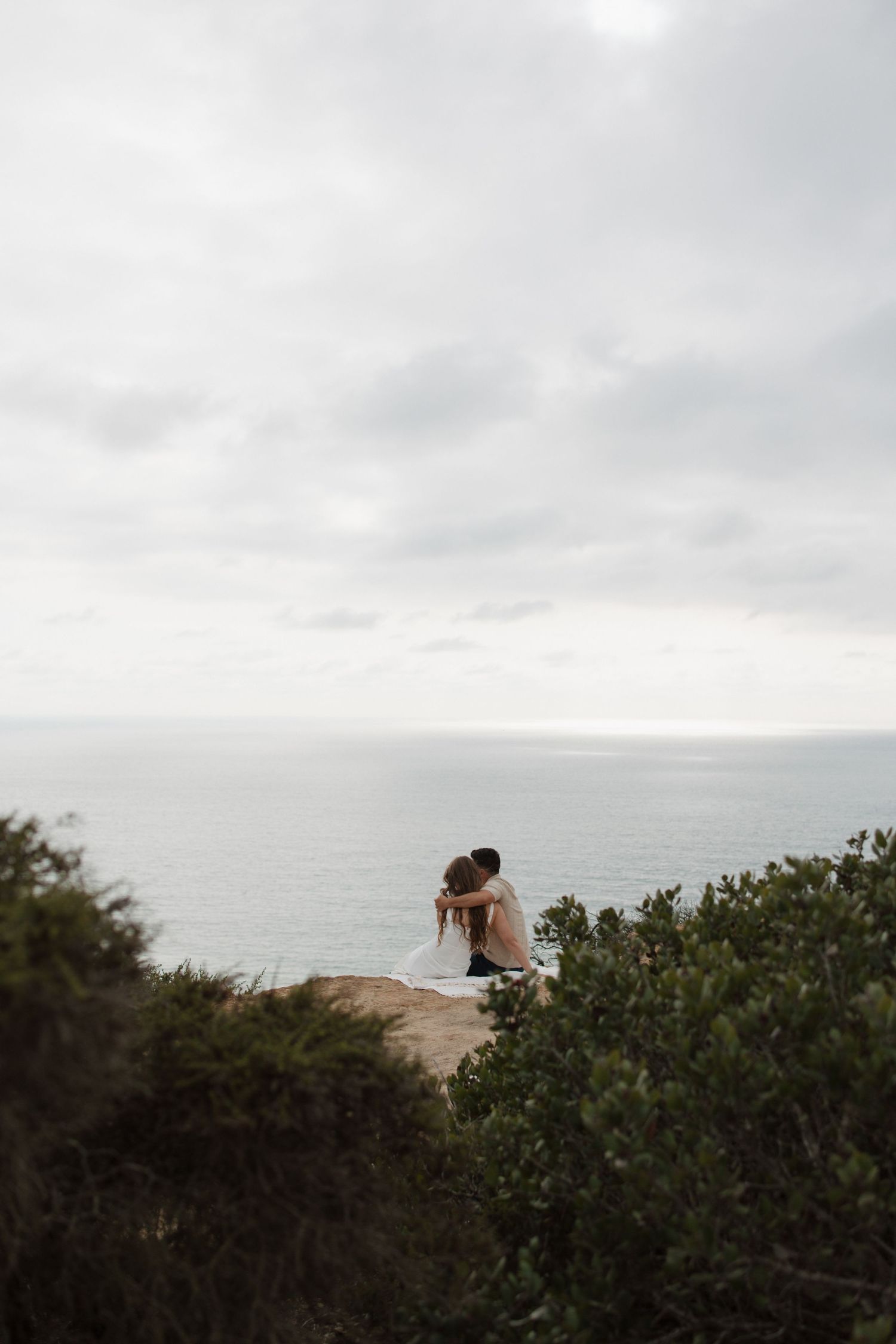 Couple kissing on a rocky cliff overlooking the vast ocean on a cloudy day.