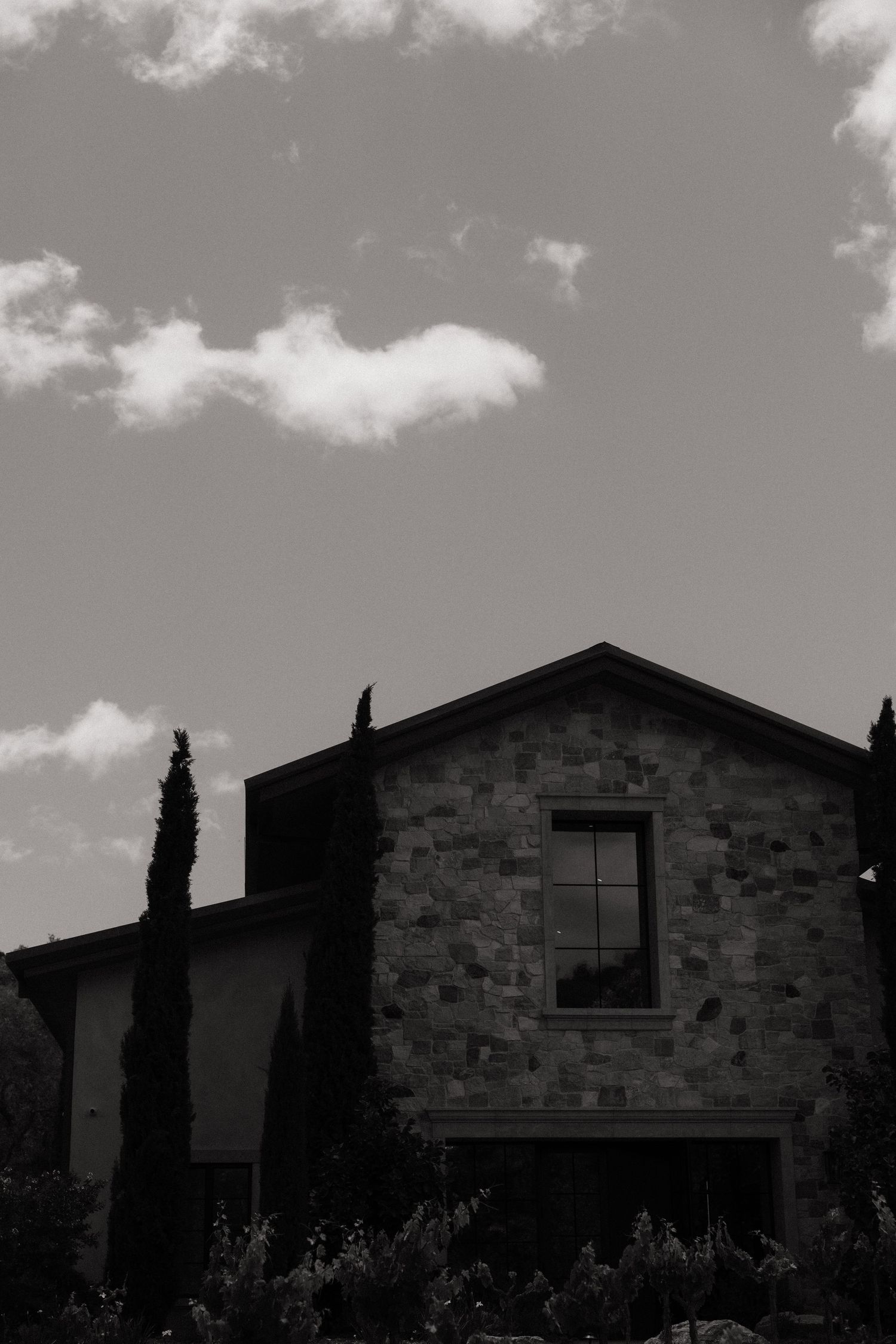 Black and white photo of a rustic stone building with cypress trees under a cloudy sky.