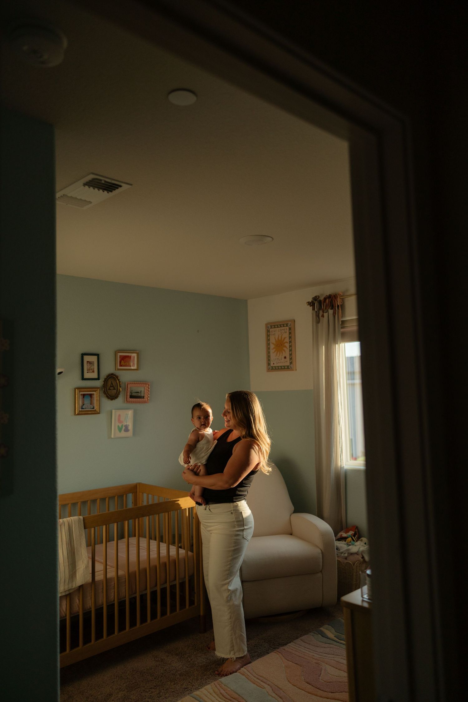 Mother holding her newborn baby in a softly lit nursery with a wooden crib and sage green walls.