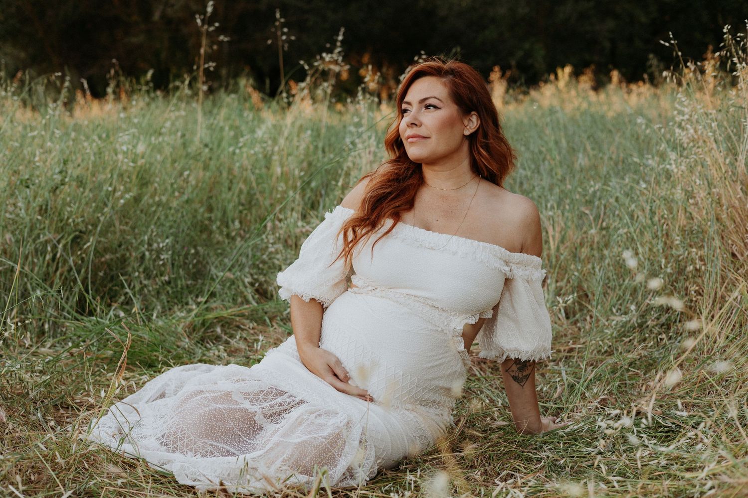 Pregnant woman in white off-shoulder gown sitting in golden meadow, maternity photography outdoors.