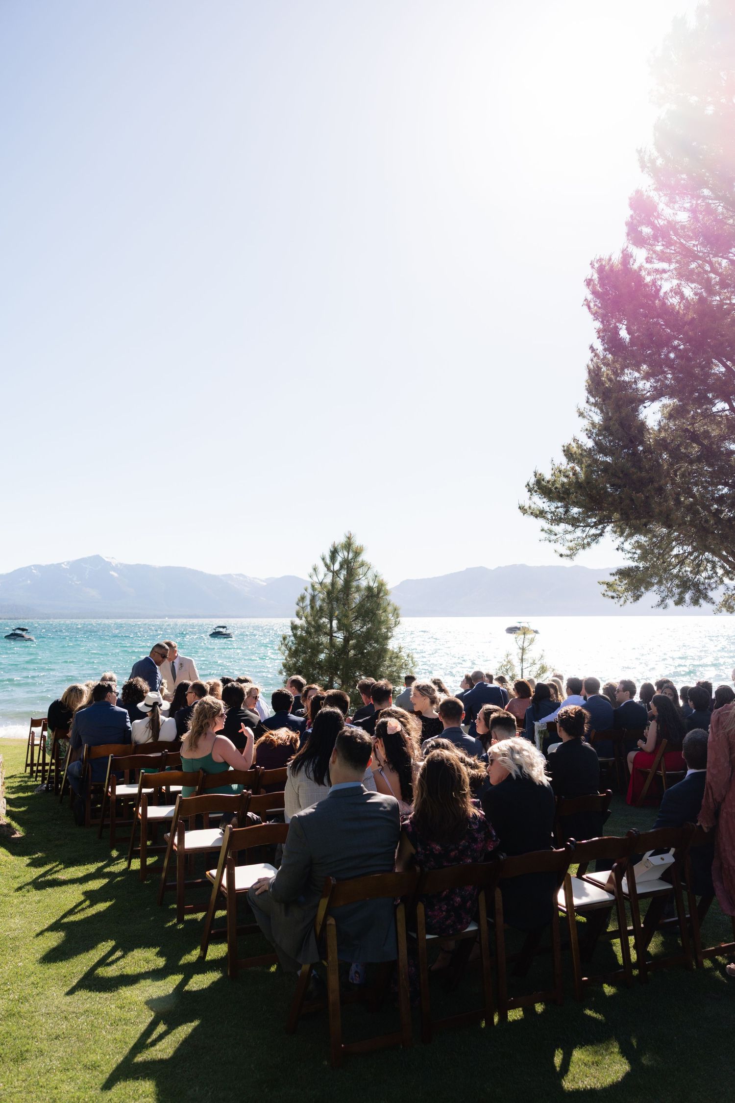 Outdoor lakeside wedding ceremony with guests seated on wooden chairs under bright sunny skies.