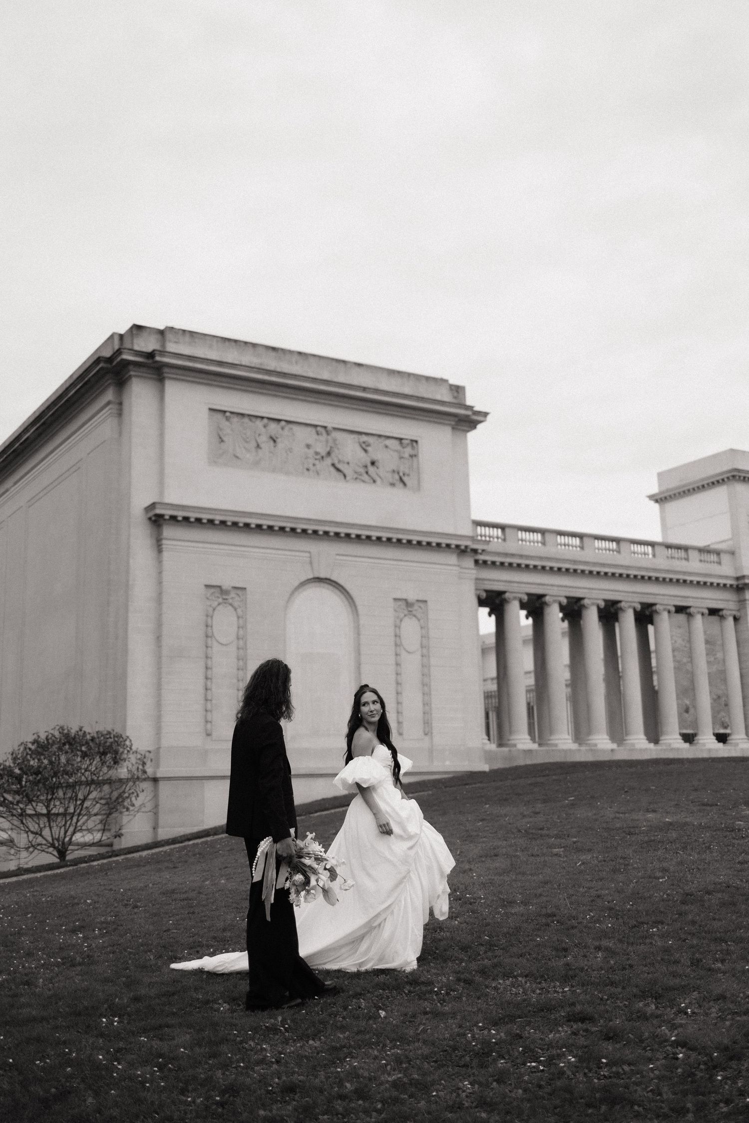 Bride and groom holding hands on green lawn before neoclassical building in black and white wedding photo.