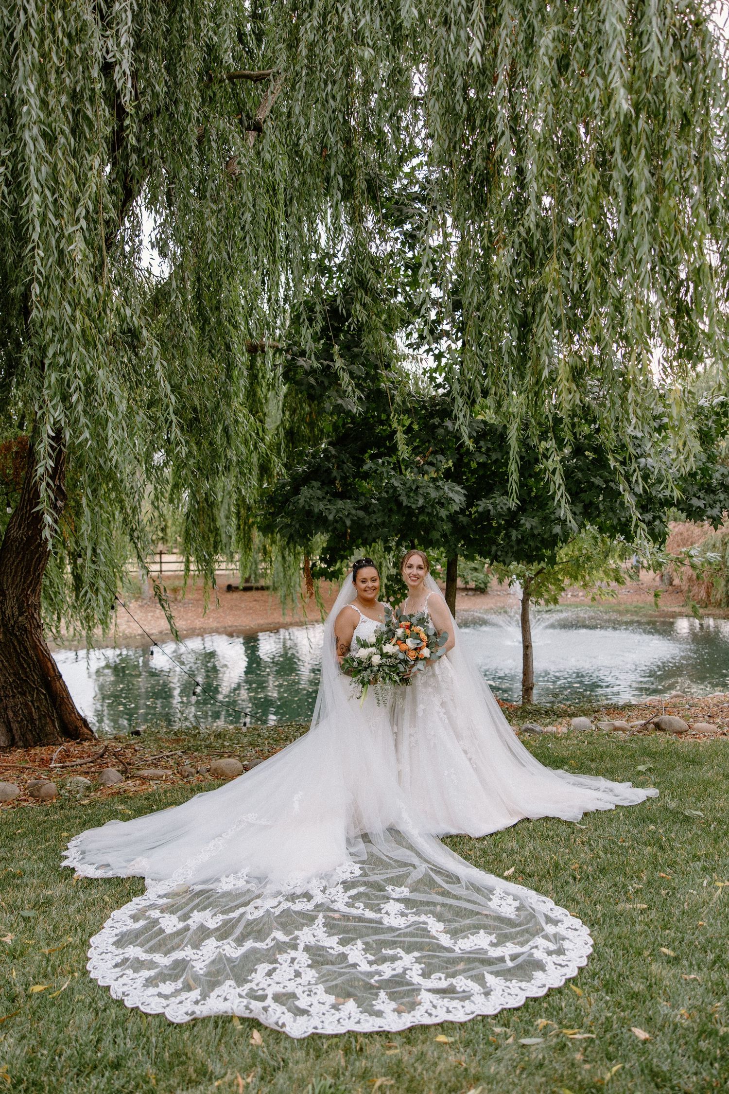 Two brides in white gowns with long lace veils posing under a weeping willow tree by a pond.