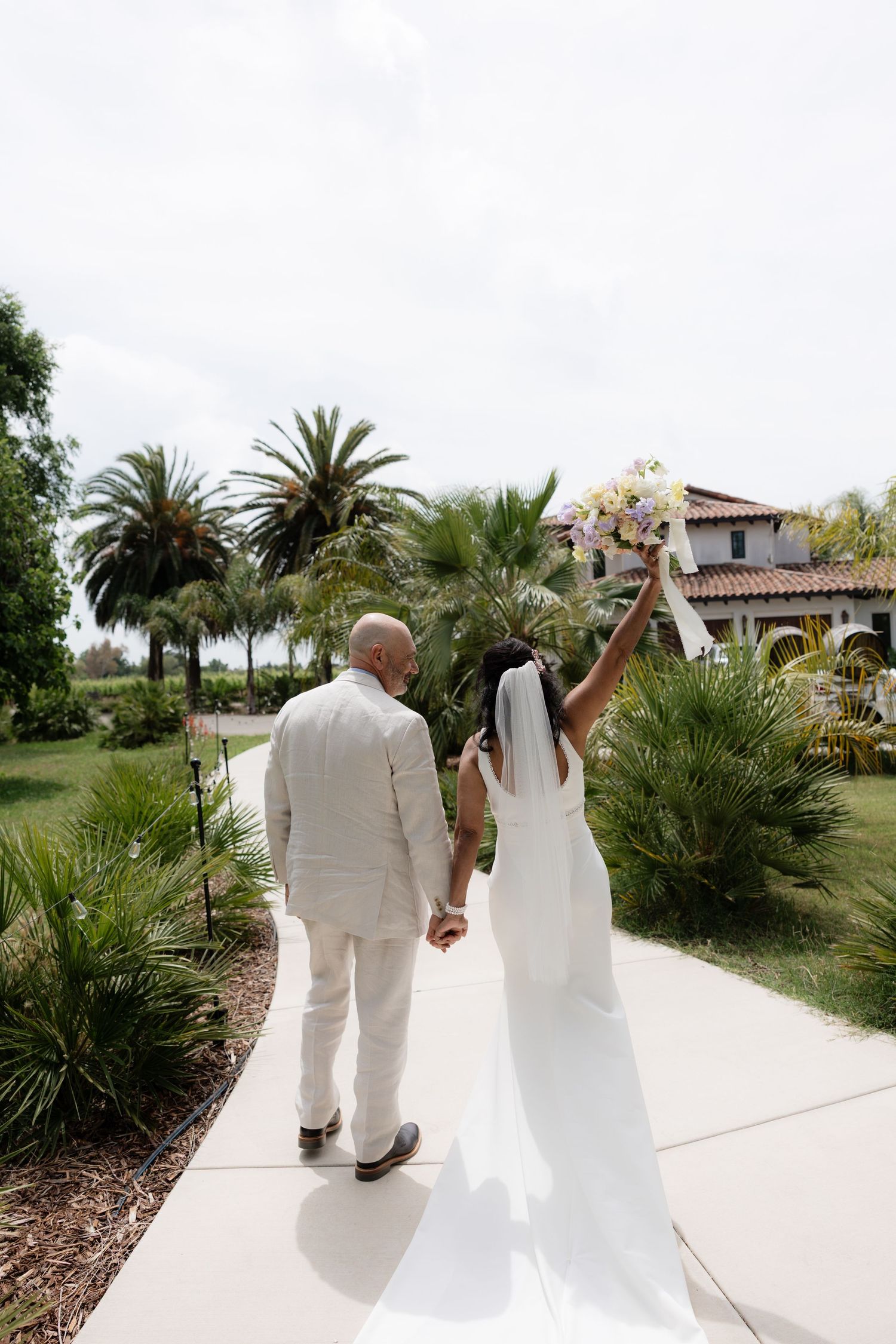 Bride raises bouquet of white and pink flowers while holding grooms hand walking toward tropical Spanish-style wedding estate.