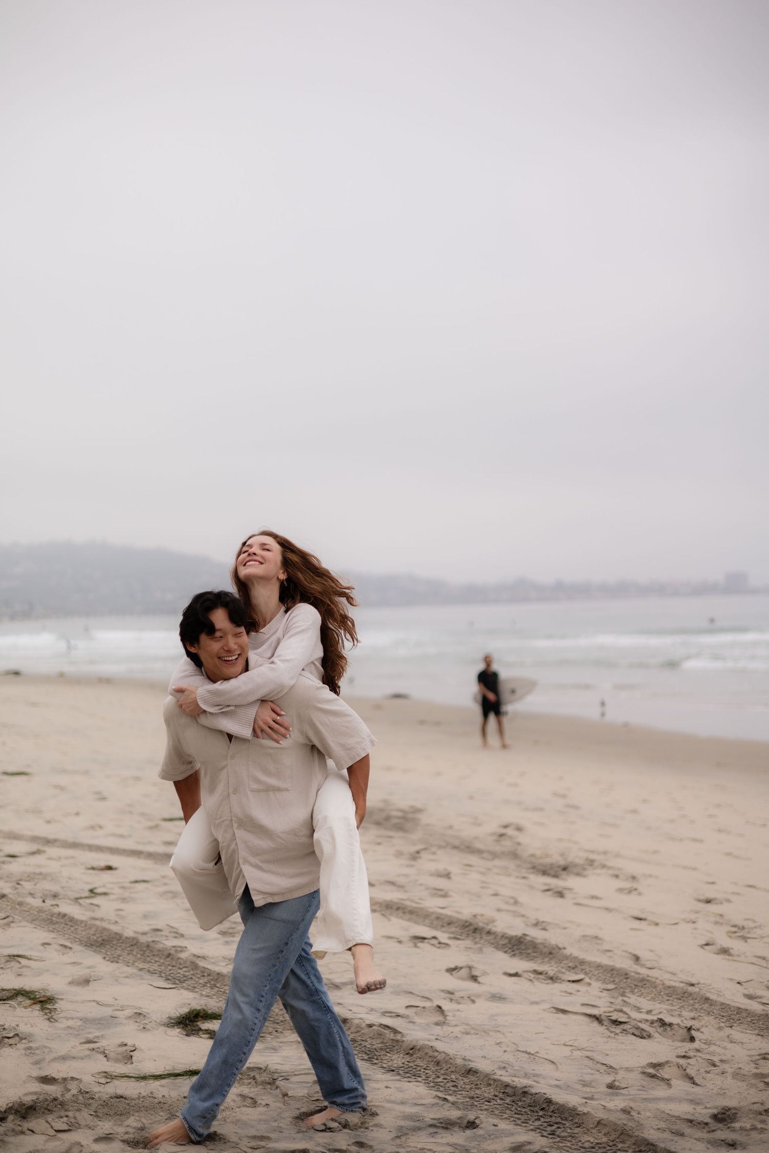 A couple enjoys a piggyback ride on a misty beach, the woman laughing joyfully as her hair blows in the wind.