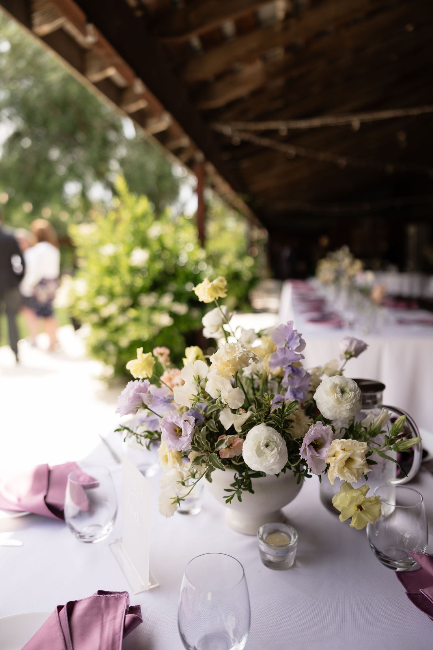 Elegant wedding reception table with pastel floral centerpiece and lavender napkins in an outdoor venue.