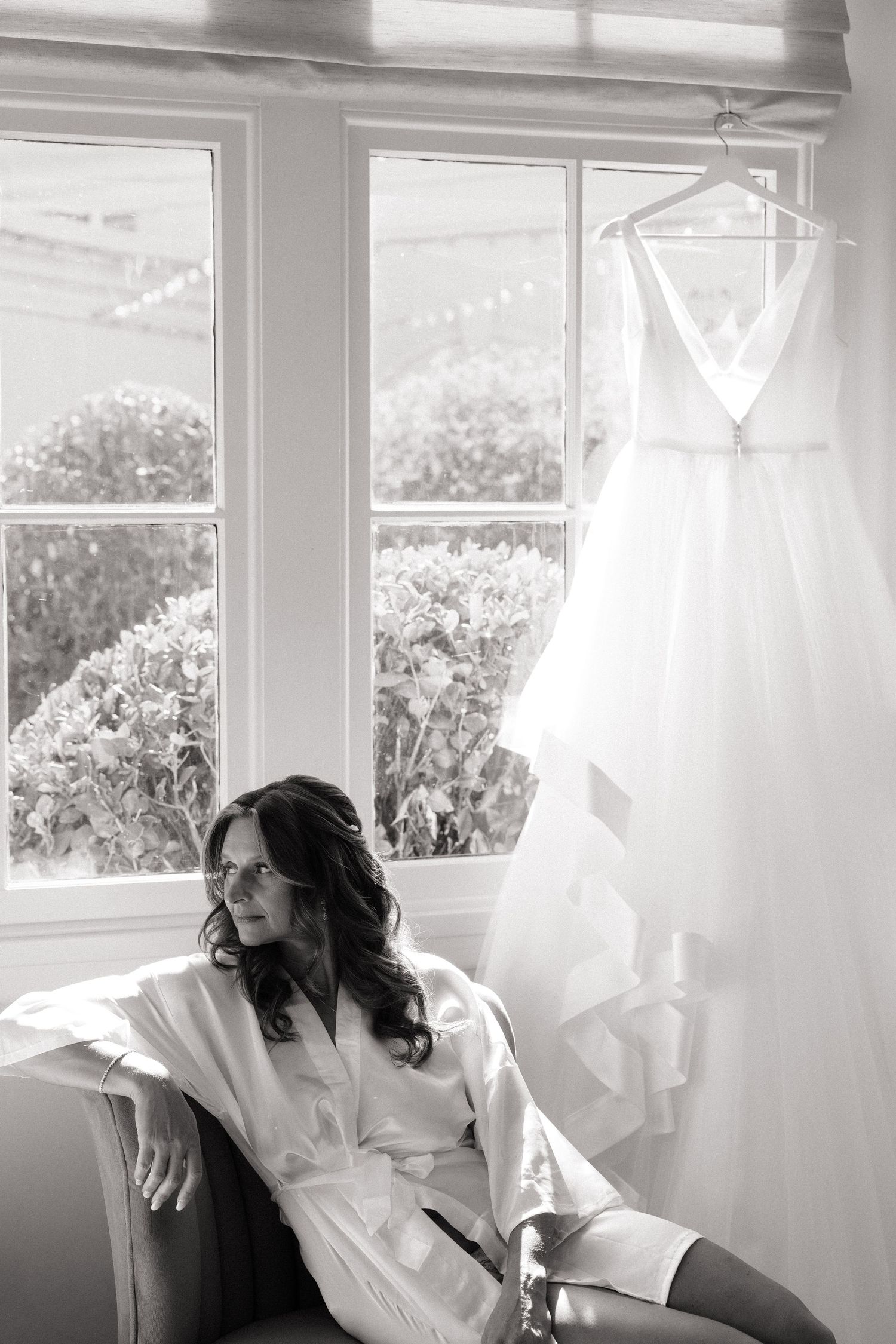 Black and white photo of a bride in a white robe sitting in a chair beside her wedding dress hanging by a window.