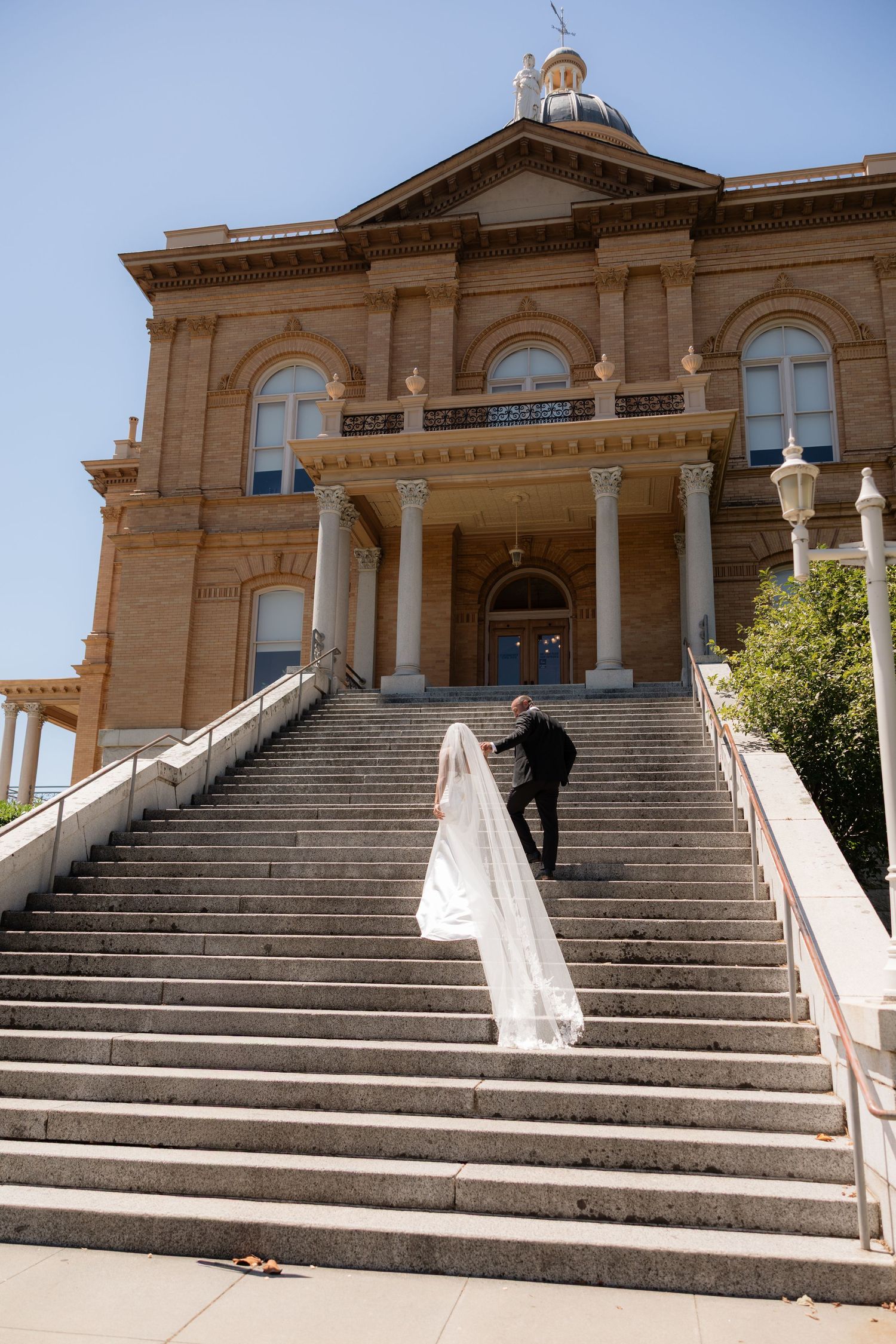Bride and groom ascending grand stone steps of historic courthouse building on their wedding day.