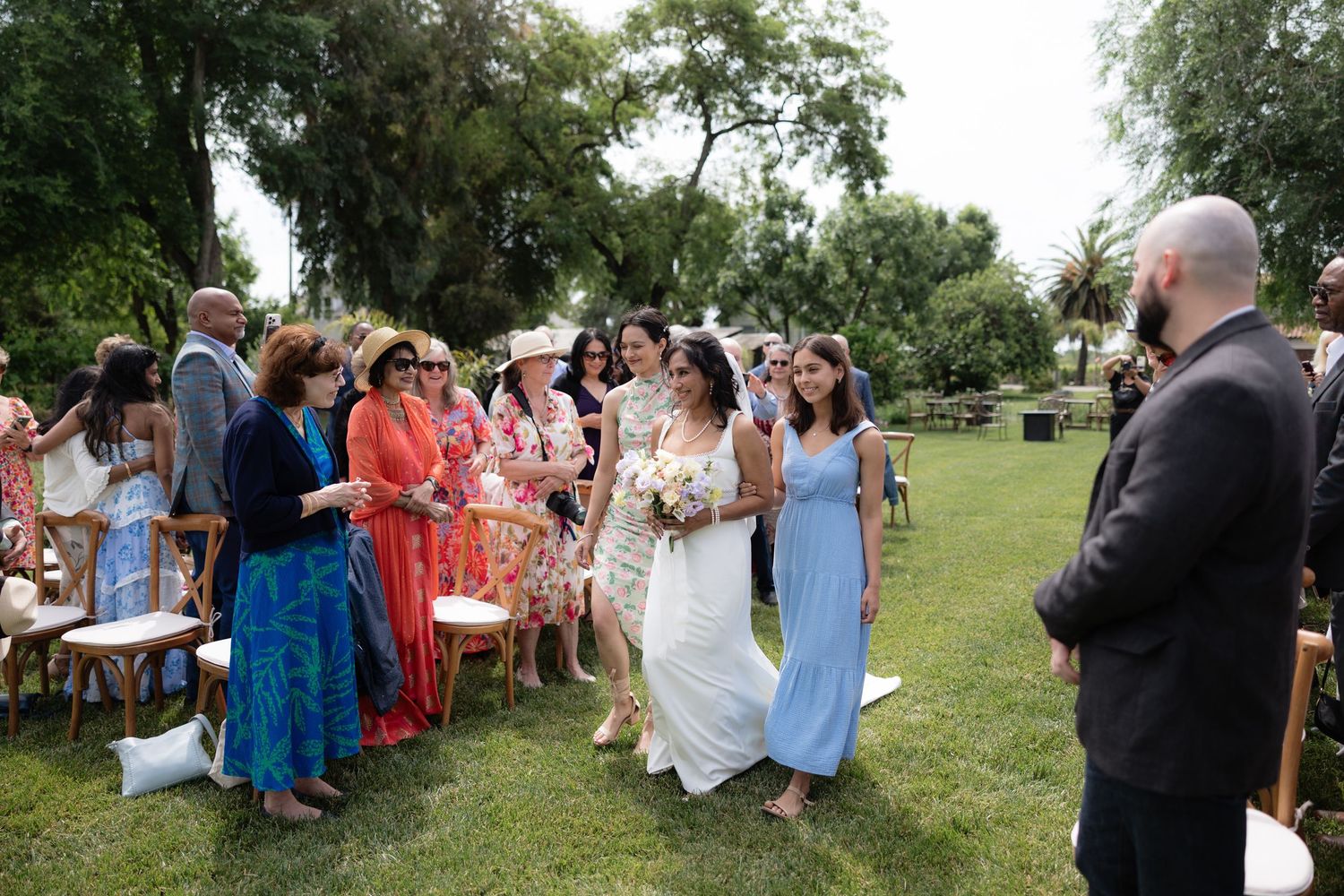 Bride walking down the outdoor aisle greeted by smiling guests during a garden wedding ceremony.