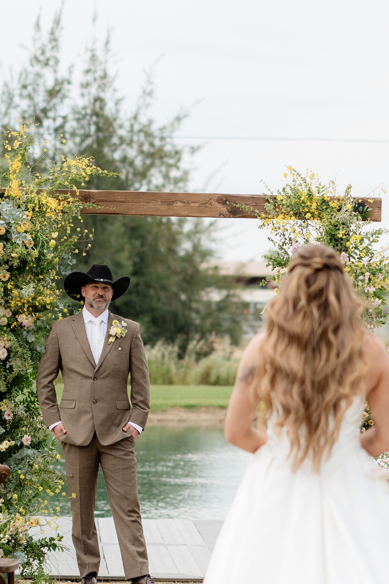 Groom in cowboy hat and tan suit watches bride approach floral wooden arch near pond.