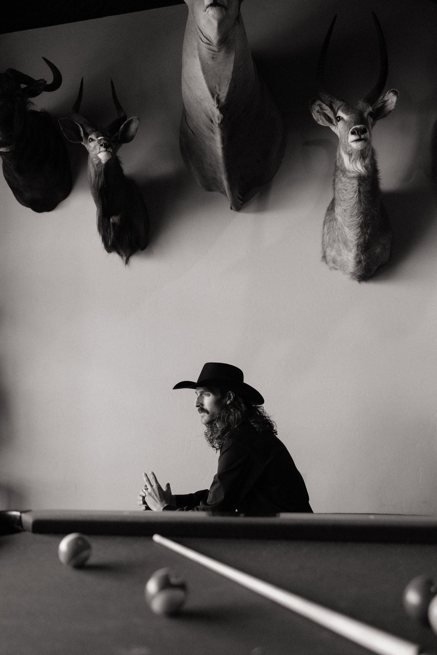 Groom in black cowboy hat sitting at a pool table beneath mounted animal trophies on the wall.